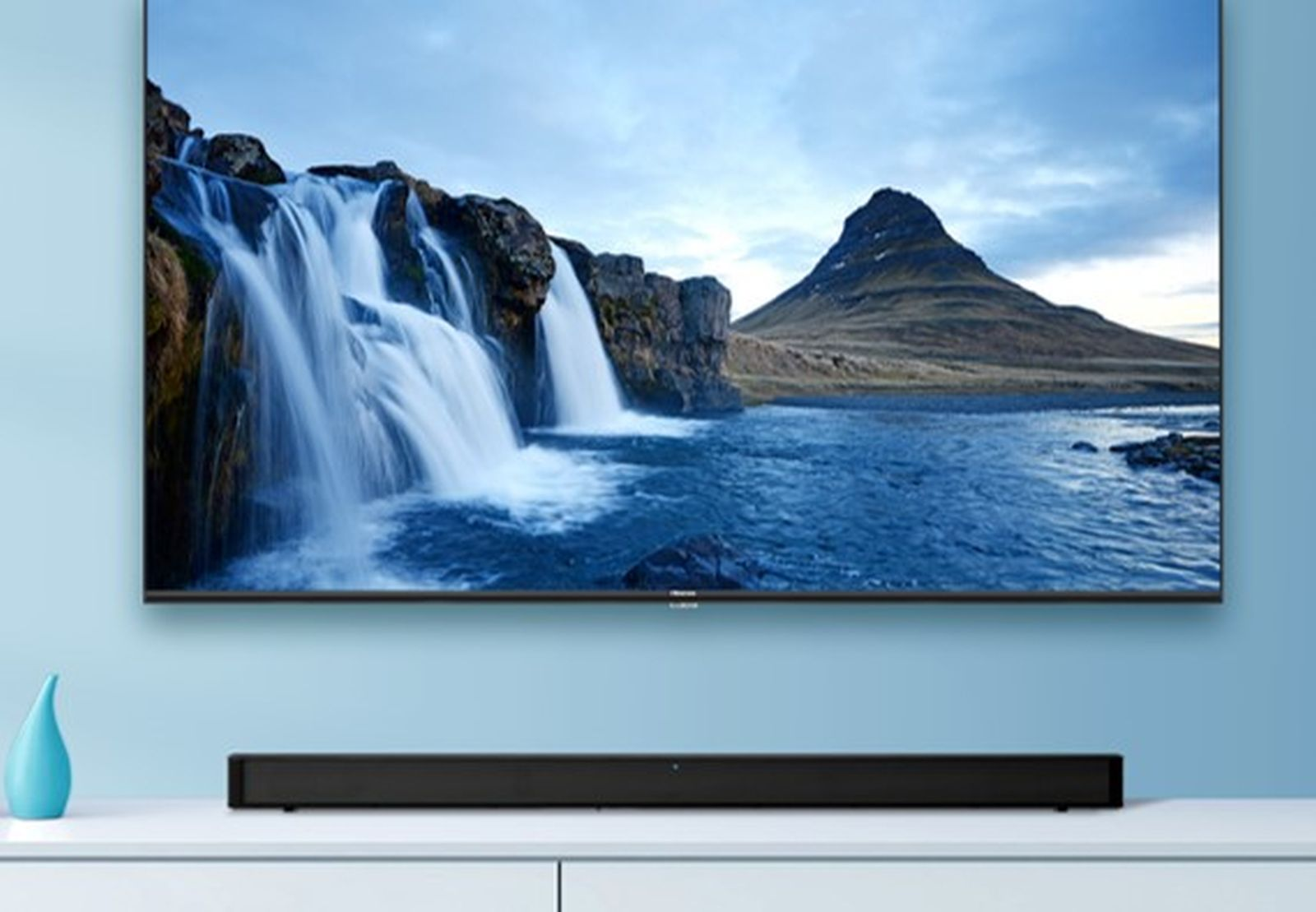Fernseher mit Wasserfall, schwarze Soundbar darunter. Blaue Wand, weißer Schrank und blaue Vase links.