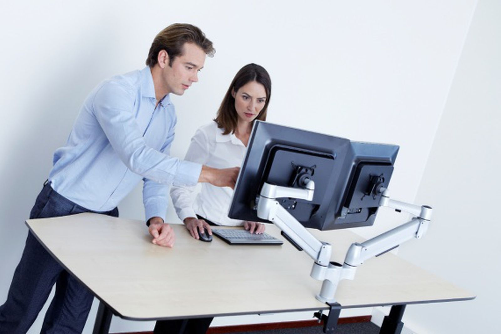 Twee mensen aan een bureau. Een man wijst naar een opstelling met twee monitoren. Een vrouw kijkt naar het scherm.