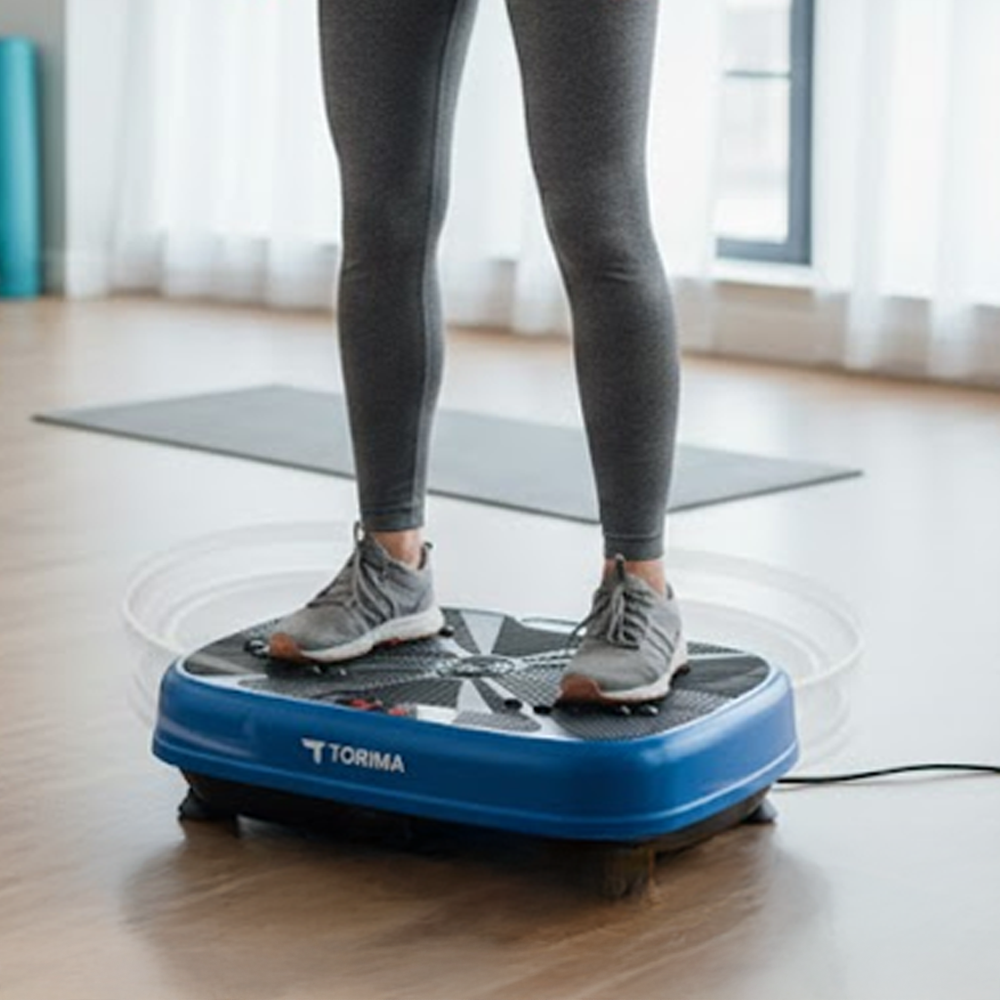 Bir kişi spor salonunda gri tayt ve spor ayakkabılarla mavi bir titreşim platformunda duruyor. Platformda Torima logosu var.