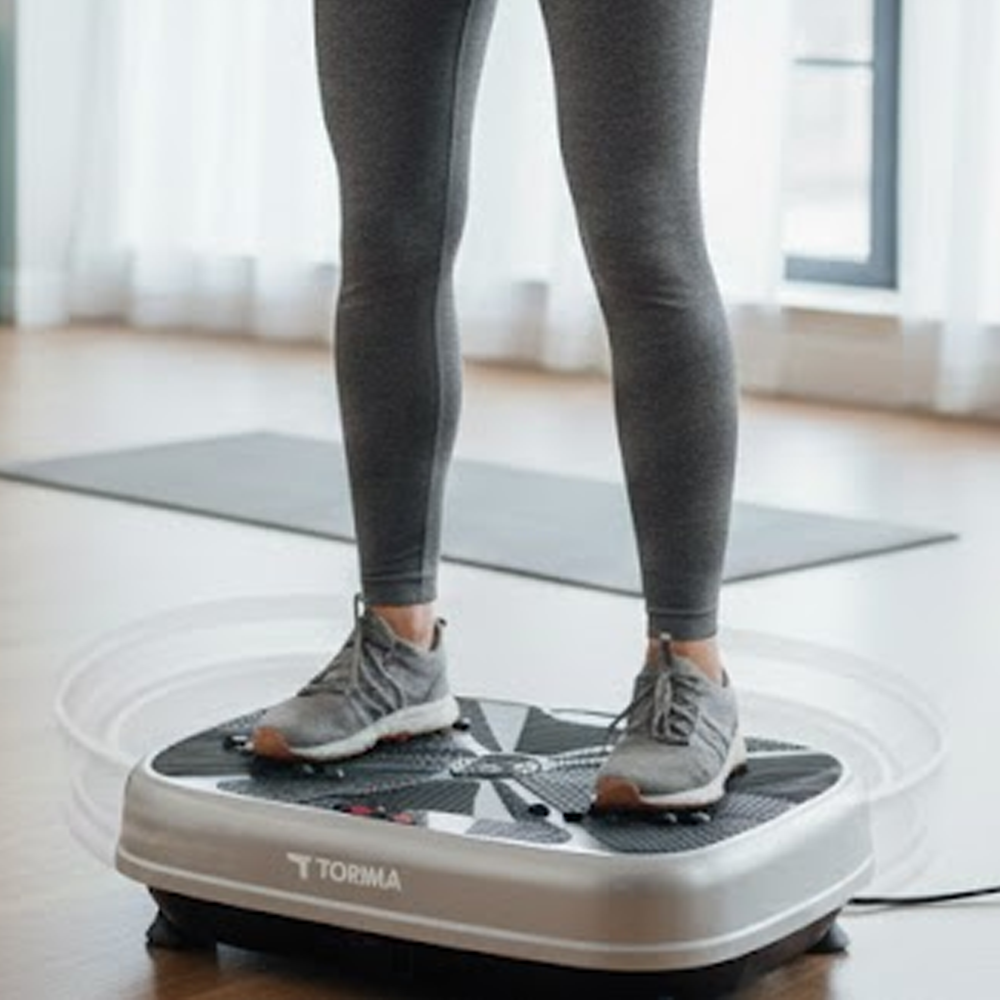 Bir kişi, gri tayt ve spor ayakkabılarla titreşimli bir egzersiz platformunda duruyor. Platform gümüş ve siyah renkte, Torima logosuyla.