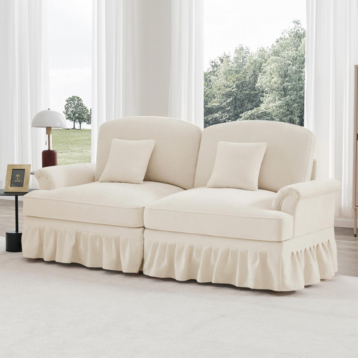 Cremefarbenes Sofa mit zwei Kissen, gerüschtem Rock und weißem Teppich. Fenster zeigt Bäume, Lampe und Bilderrahmen auf einem Beistelltisch.