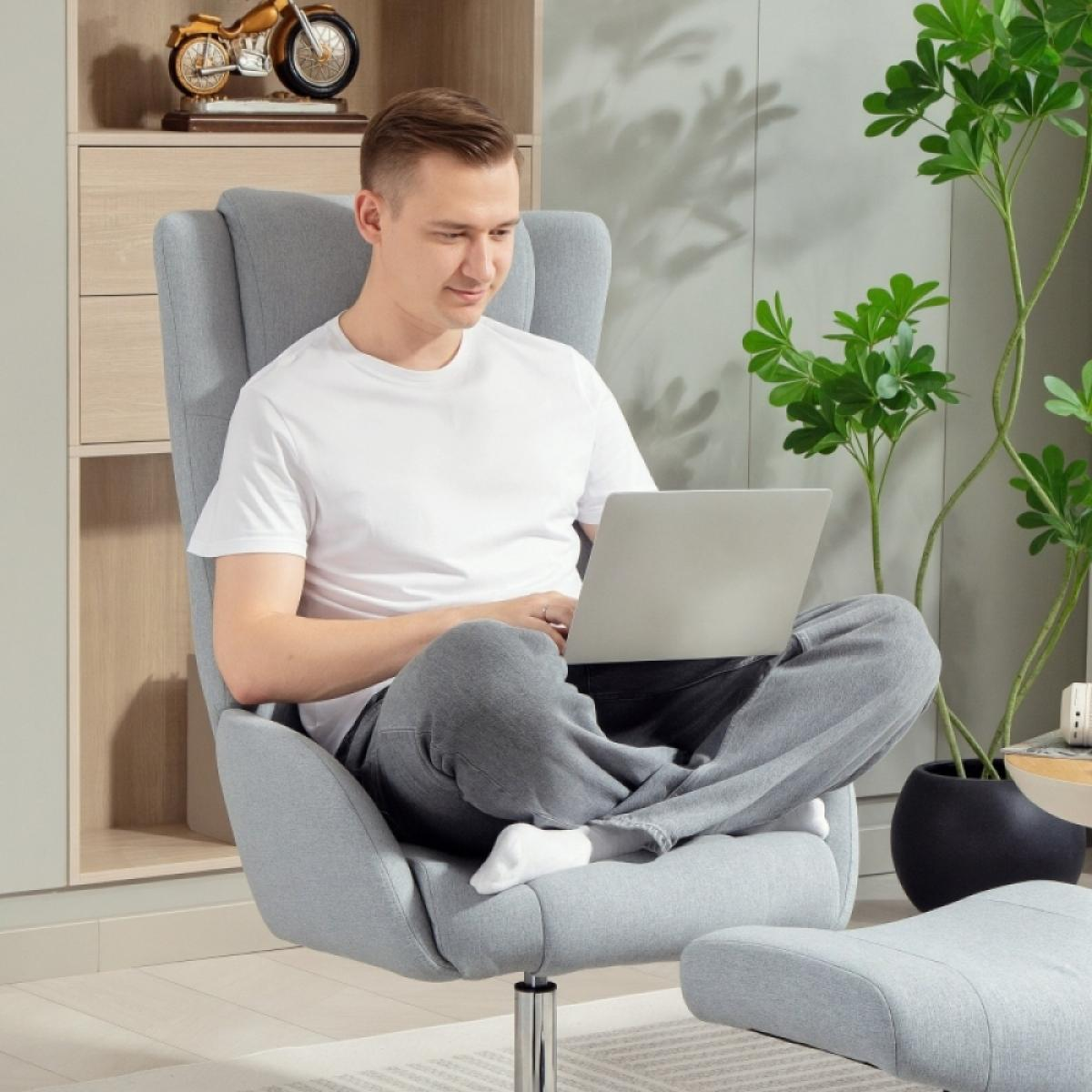 Ein Mann sitzt in einem grauen Stuhl und benutzt einen Laptop. Er trägt ein weißes Hemd und graue Hose. Grüne Pflanzen und ein Modellmotorrad sind sichtbar.
