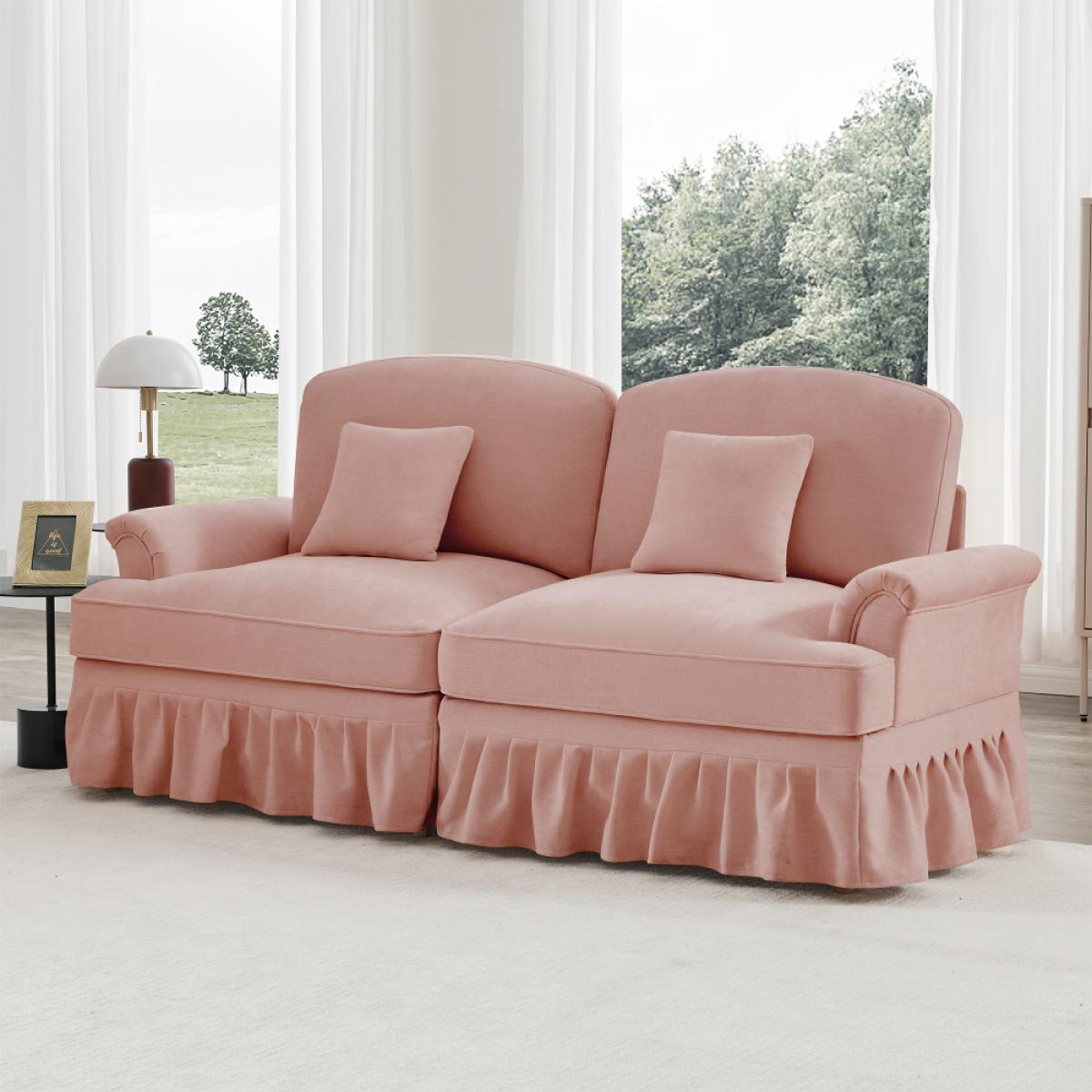 Ein rosa Sofa mit zwei Kissen steht vor einem Fenster. Daneben ein kleiner Tisch mit Lampe und Bilderrahmen.
