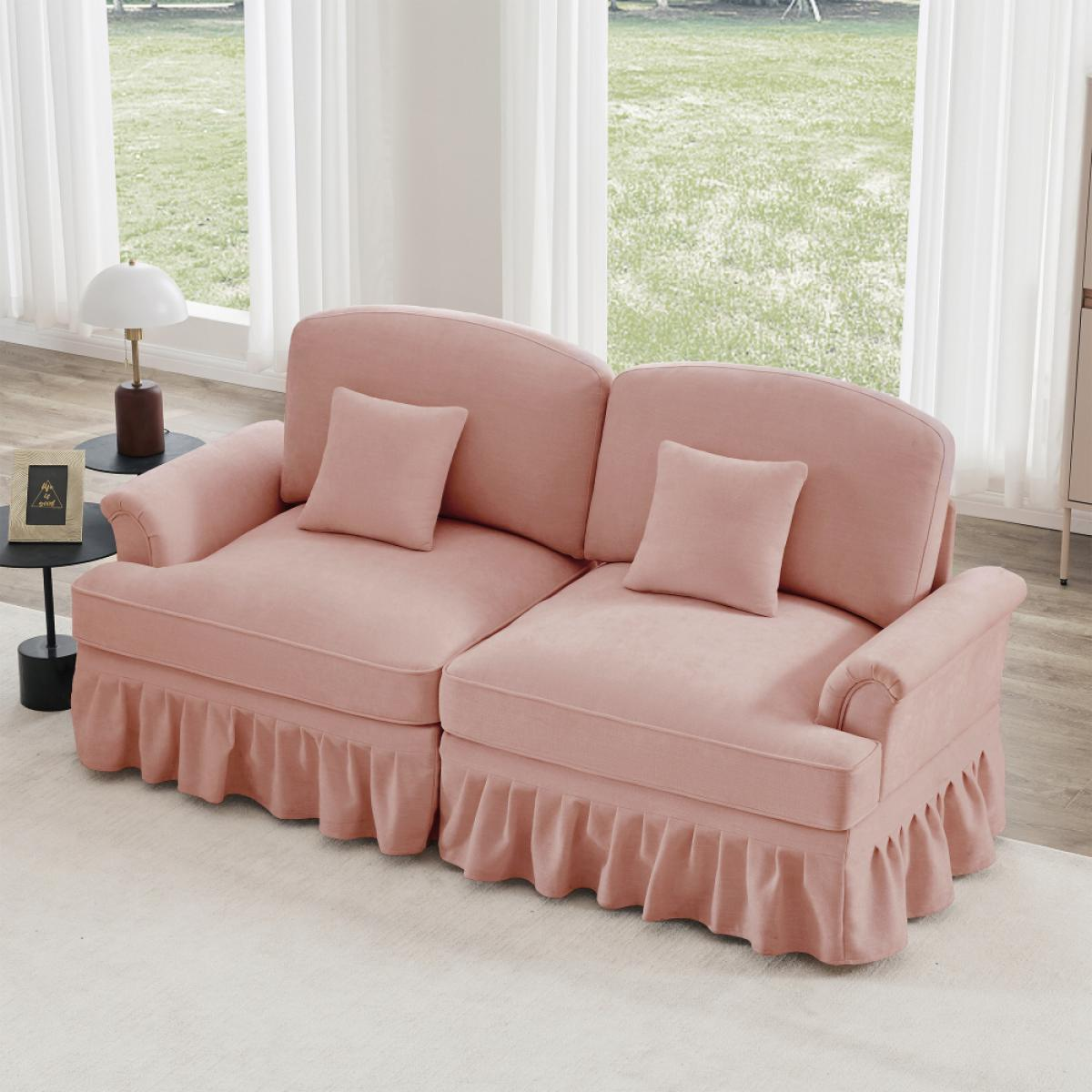 Ein rosafarbenes Sofa mit zwei Abschnitten, zwei Kissen und einem Rock. Ein kleiner Tisch mit Lampe und Bilderrahmen. Ein Fenster mit weißen Vorhängen.