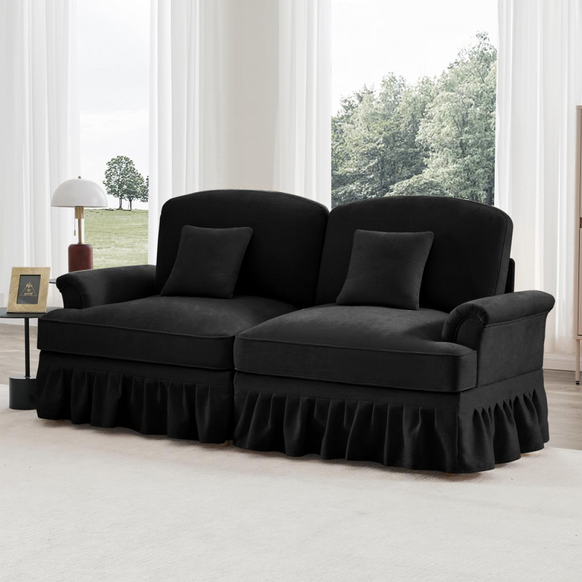 Ein schwarzes Sofa mit zwei Kissen steht auf einem weißen Teppich. Ein Beistelltisch mit Lampe und ein Fenster mit Bäumen im Hintergrund.