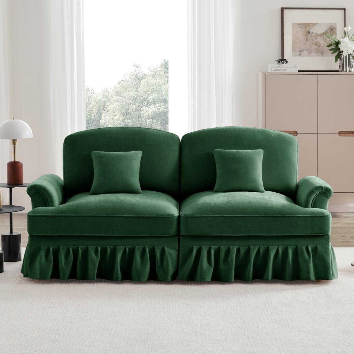 Ein grünes Sofa mit zwei Kissen. Es hat einen gerüschten Sockel und steht auf einem cremefarbenen Teppich. Eine Lampe und ein Schrank sind sichtbar.