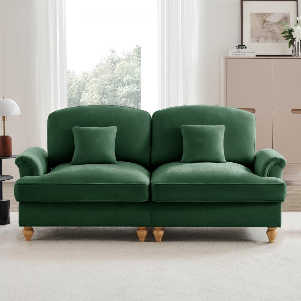Ein grünes Samtsofa mit zwei Kissen steht auf einem hellen Teppich. Hinter dem Sofa befindet sich ein Fenster mit weißen Vorhängen.