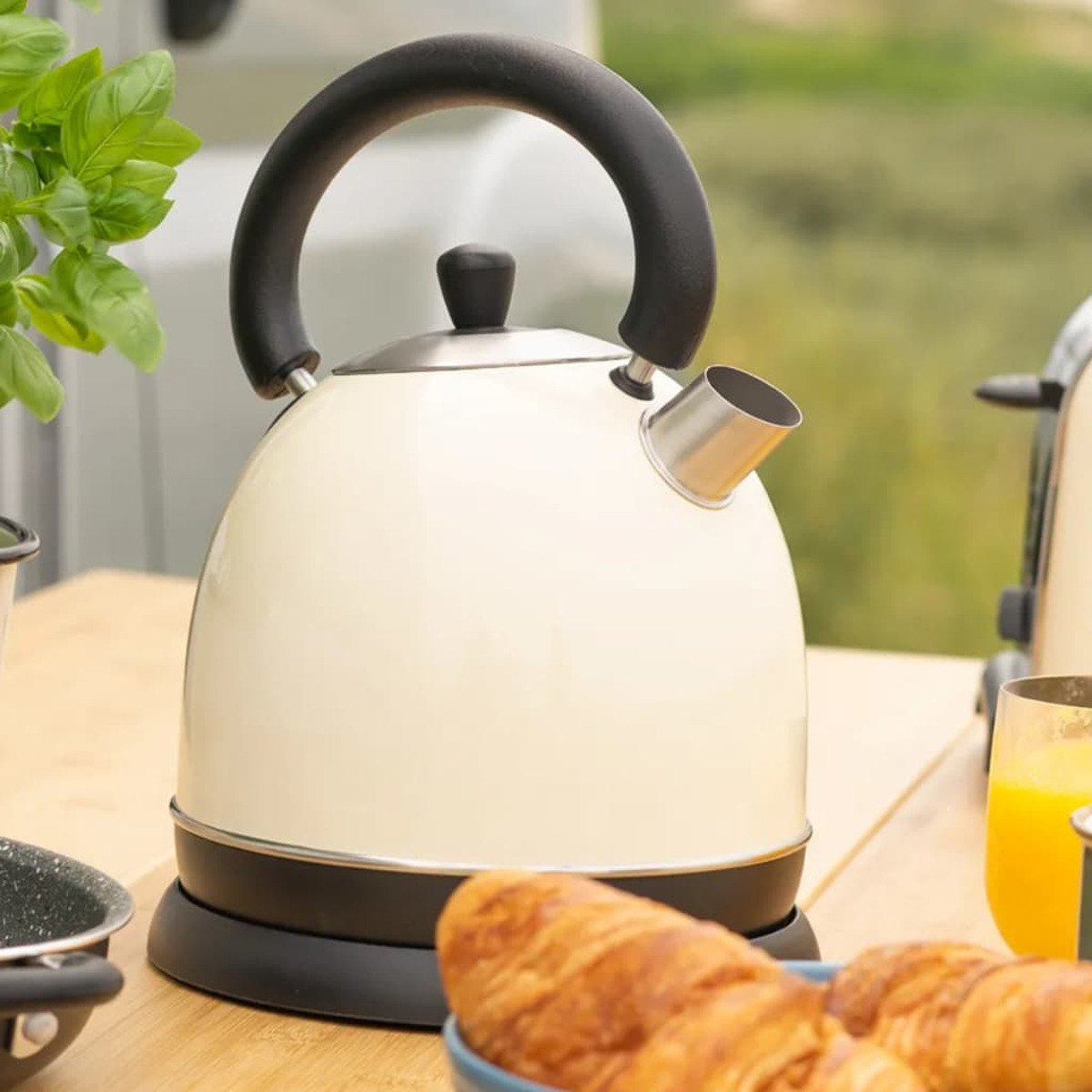 Un bollitore color crema con manico nero su una superficie di legno con croissant e bicchiere di succo.
