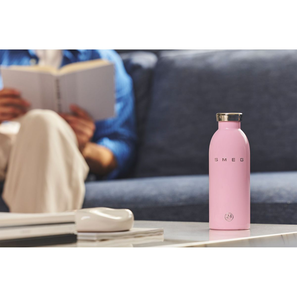 Pembe bir Smeg su şişesi masada duruyor. Arka planda bir kişi kanepede kitap okuyor. Yakınlarda beyaz bir tabak var.