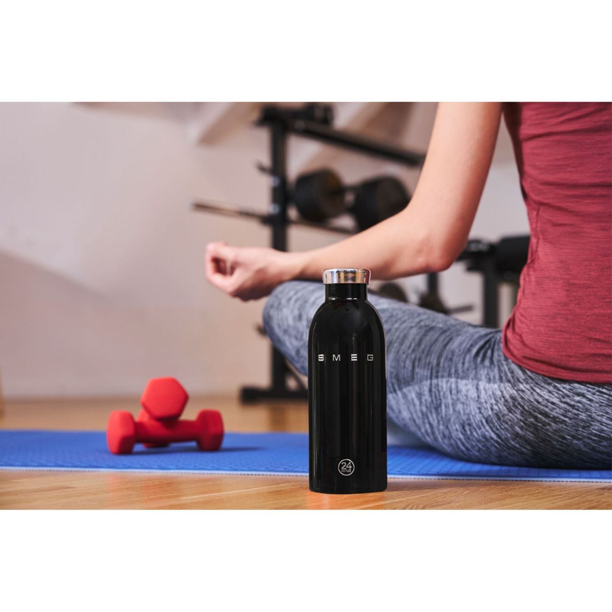Bir kişi siyah bir su şişesi ve kırmızı bir dambılın yanında meditasyon yapıyor. Sahne bir spor salonunda.