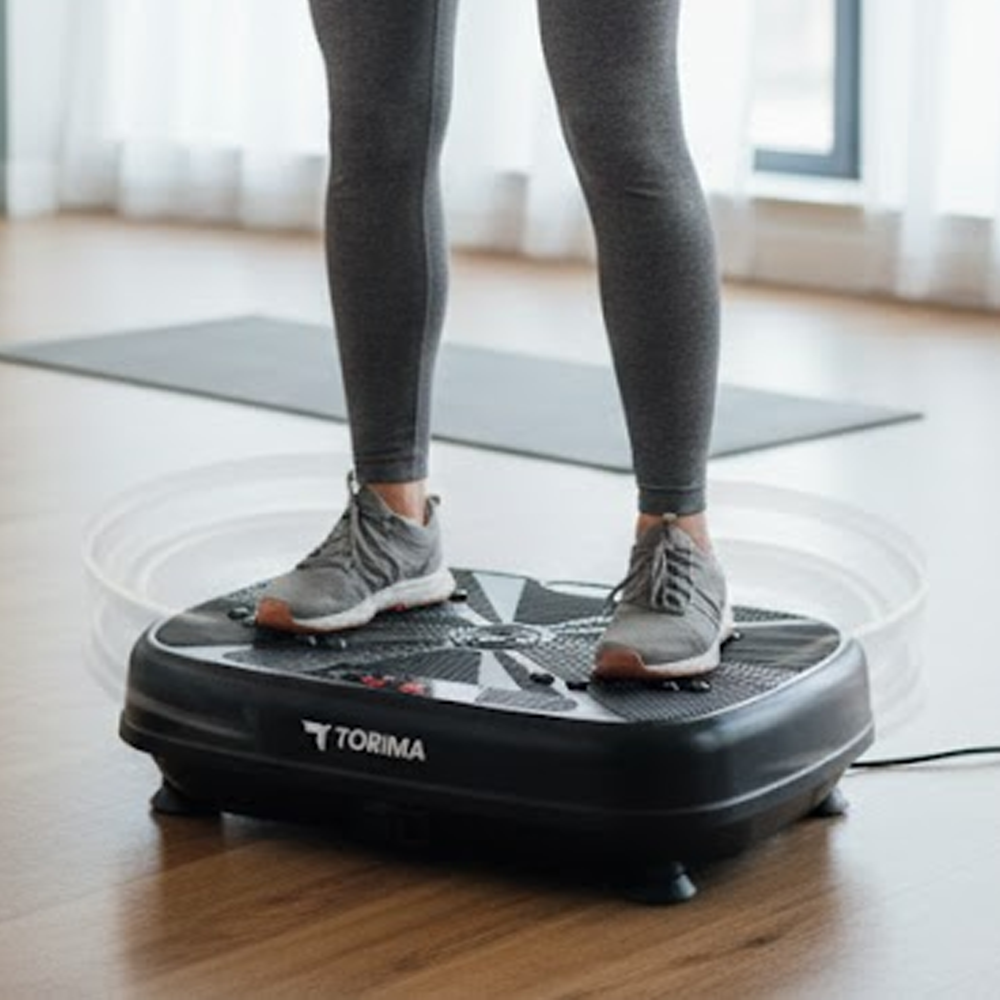 Siyah bir titreşim platformunda duran kişi. Kişi gri tayt ve ayakkabı giyiyor. Arka planda bir yoga matı var.