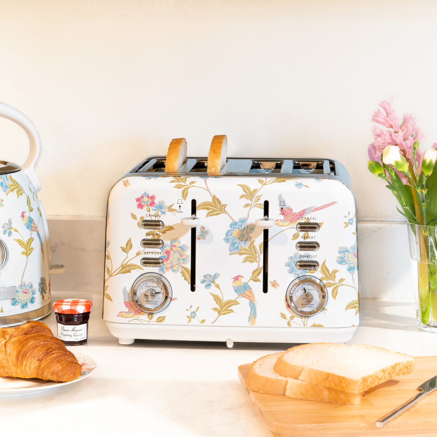 Ein floraler Toaster mit Brot, ein Wasserkocher, Croissant, Marmelade, Blumen und ein Schneidebrett mit Brotscheiben. Die Szene ist hell.