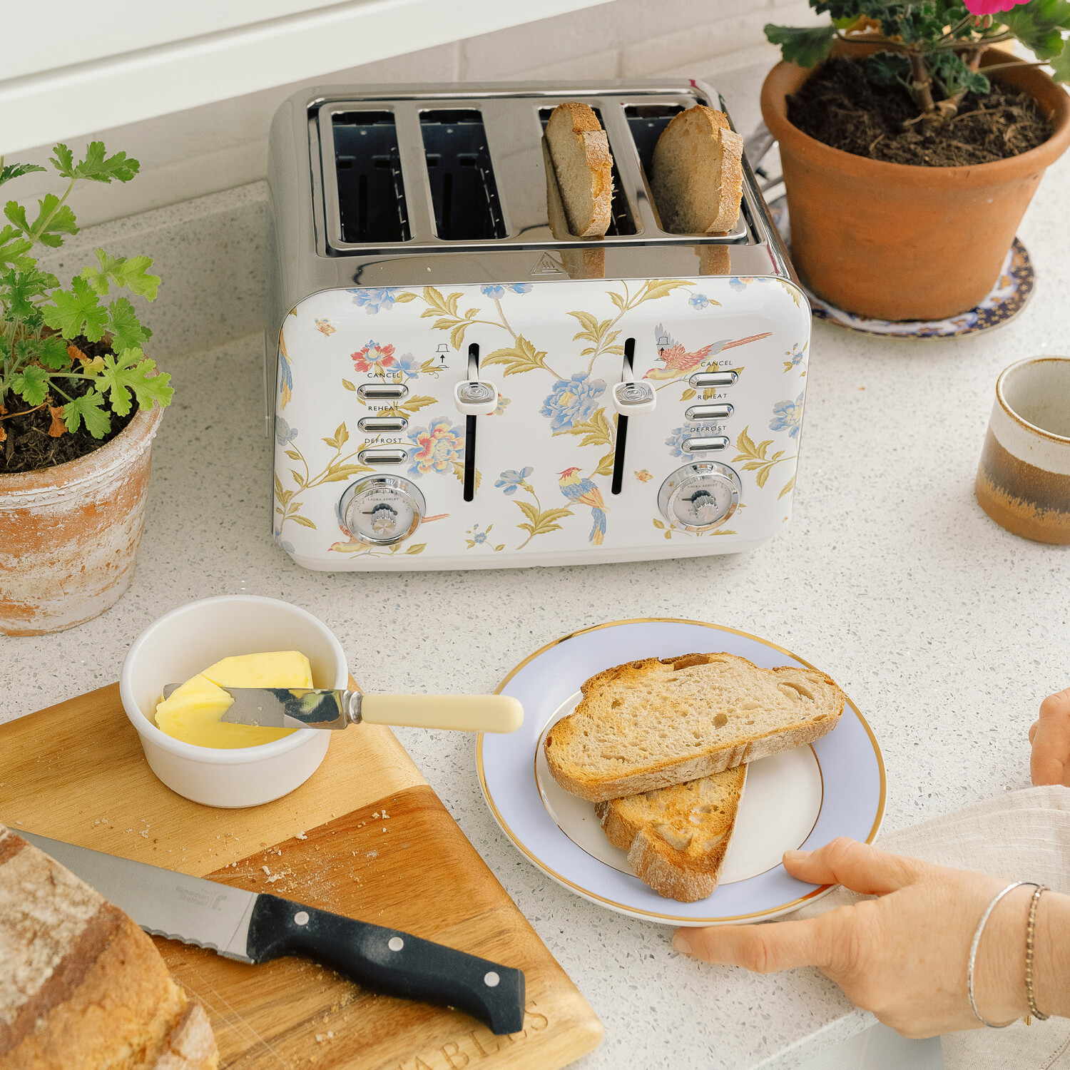 Ein Toaster mit Blumenmuster mit Toast, Butter, Messer, Brot und einem Teller mit Toast auf einer Arbeitsplatte. Frauenhand mit Armbändern.
