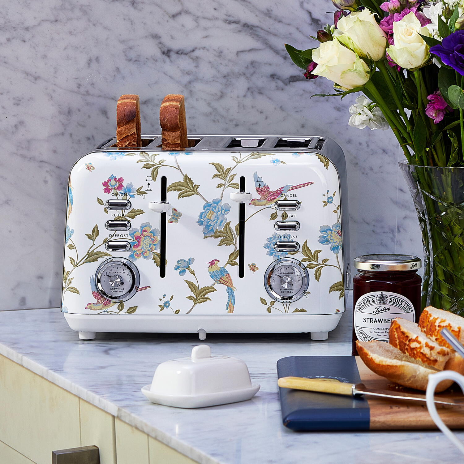 Ein floraler Toaster mit getoastetem Brot, Blumen, Marmelade, Butterdose und Brot auf einer Arbeitsplatte.