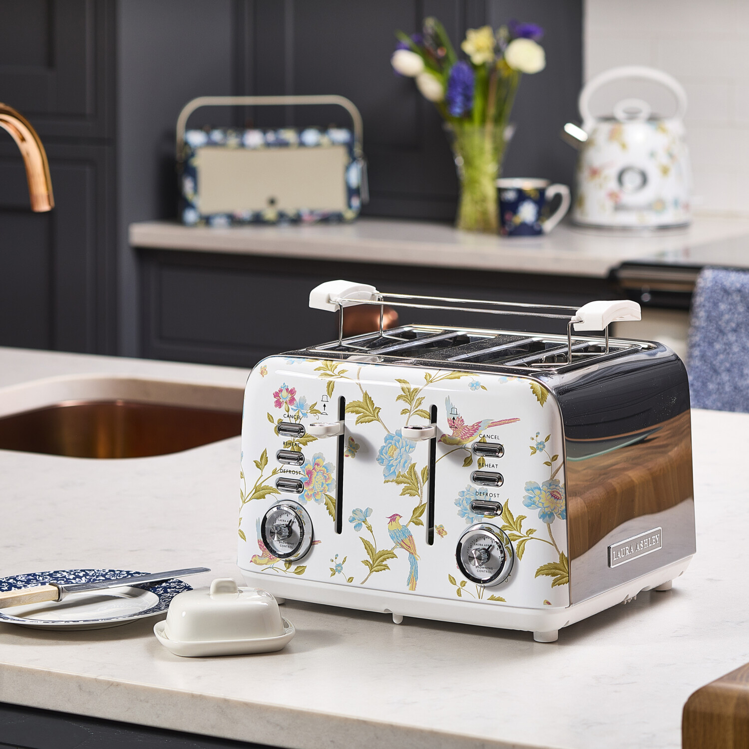 Ein floraler Toaster, eine Butterdose, ein Teller mit Messer, ein Wasserkocher, eine Tasse und Blumen stehen auf einer Arbeitsplatte. Küchenhintergrund.