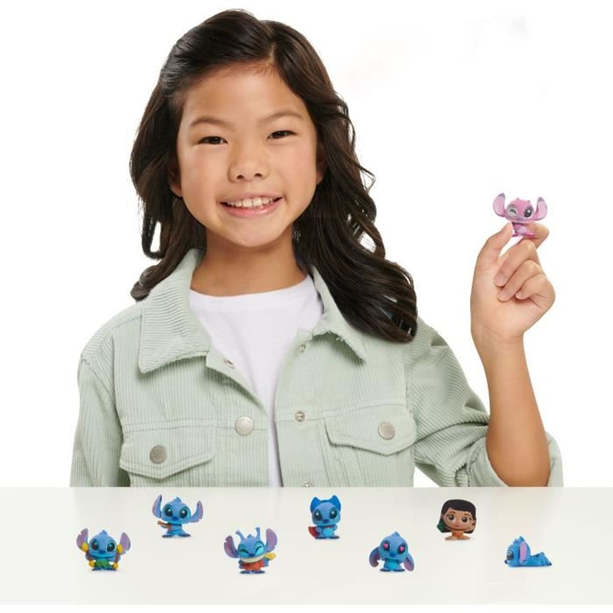 Una niña sonríe, sosteniendo una figura rosa de Stitch. Otras figuras de Stitch están en una superficie blanca. La niña lleva una chaqueta verde.