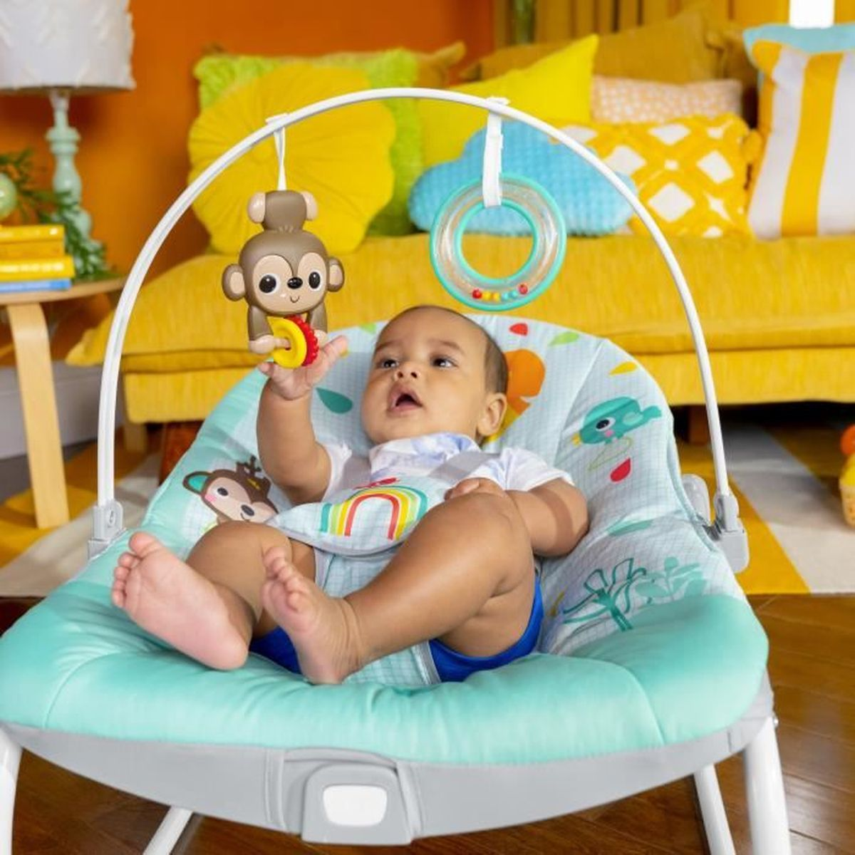 Ein Baby sitzt in einer blauen Wippe und blickt auf ein Affenspielzeug. Gelbe Kissen und eine Couch sind im Hintergrund.