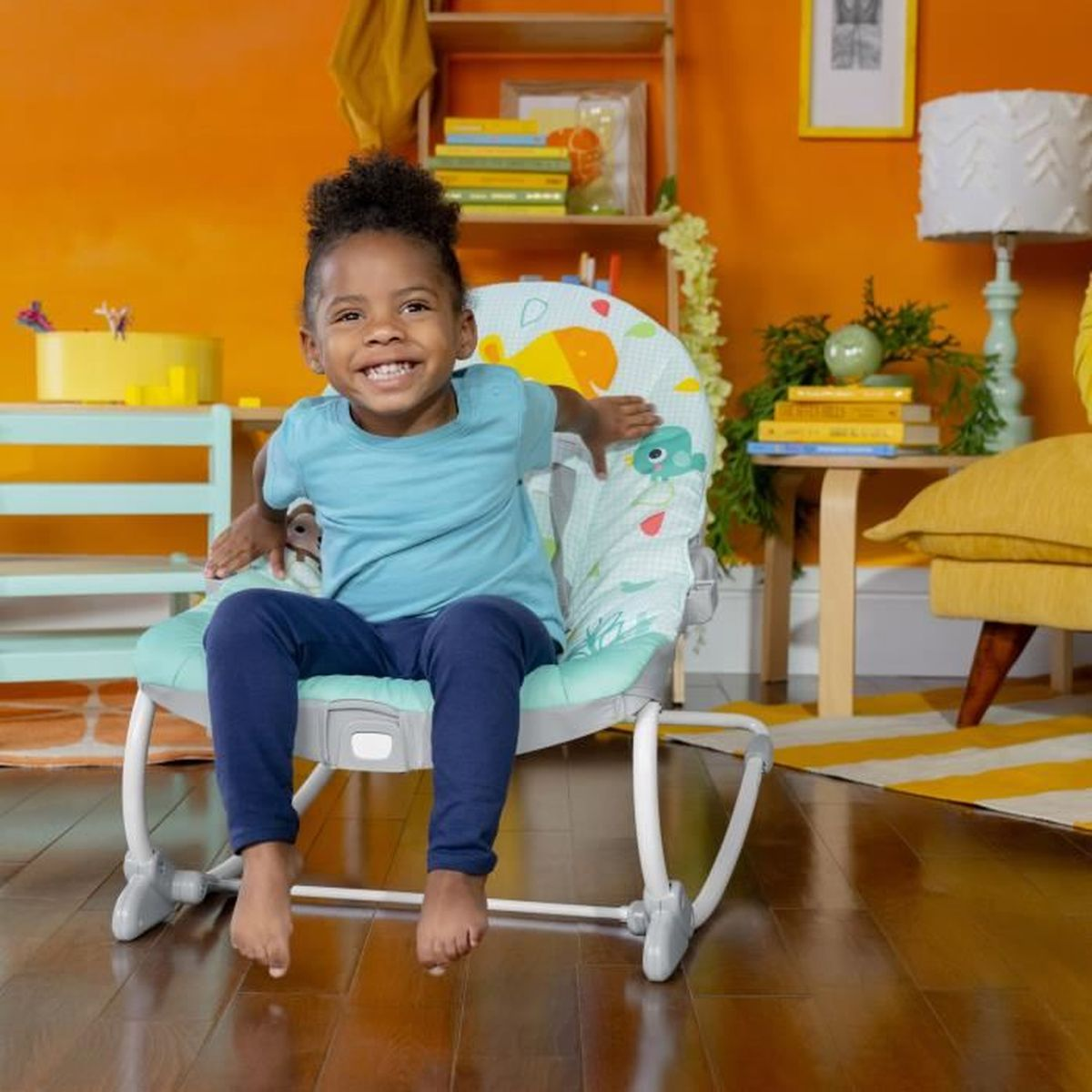 Ein lächelndes Kind sitzt in einer Babywippe. Der Hintergrund umfasst orangefarbene Wände, ein gelbes Sofa und einen gestreiften Teppich.