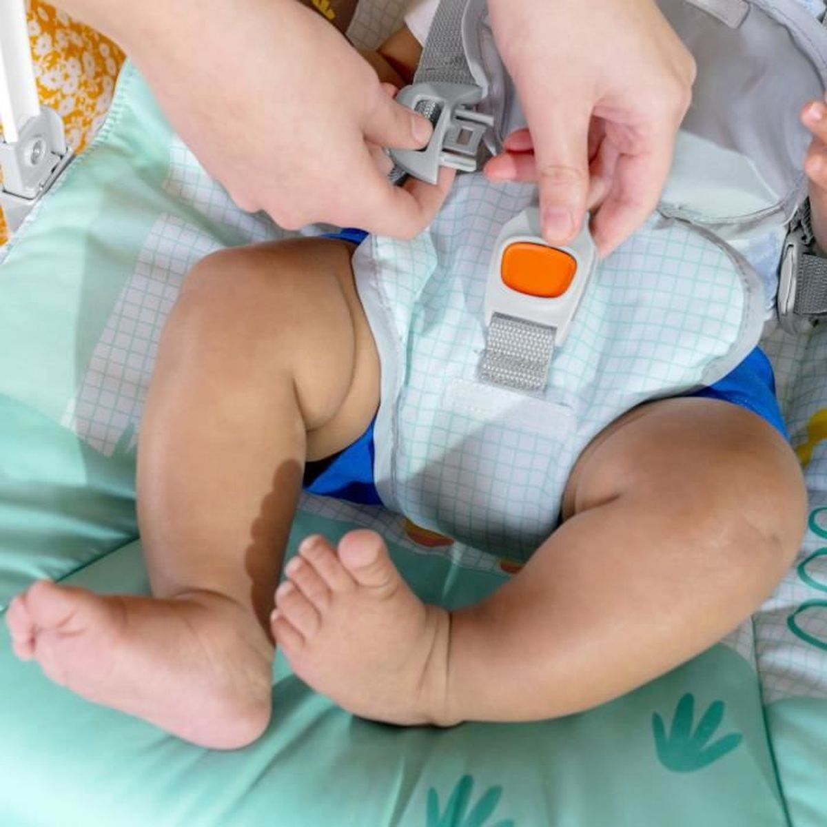 Ein Baby in einem Sitz, Hände befestigen Gurte. Der Sitz ist grün, das Baby trägt blaue Shorts. Füße sichtbar.