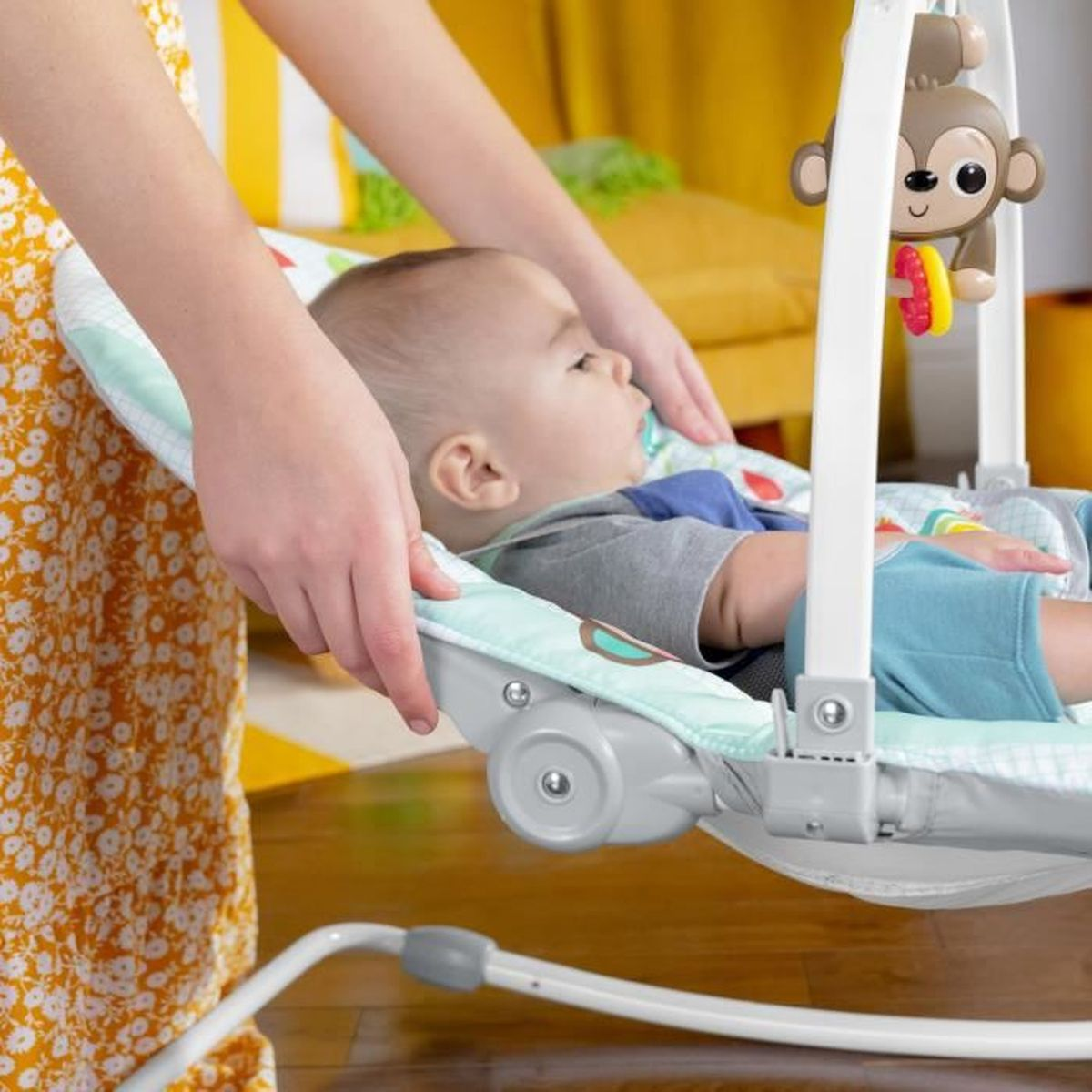 Ein Baby liegt in einer Babywippe. Eine Person hält die Wippe. Ein Spielzeugaffe hängt über dem Baby. Der Hintergrund ist gelb.