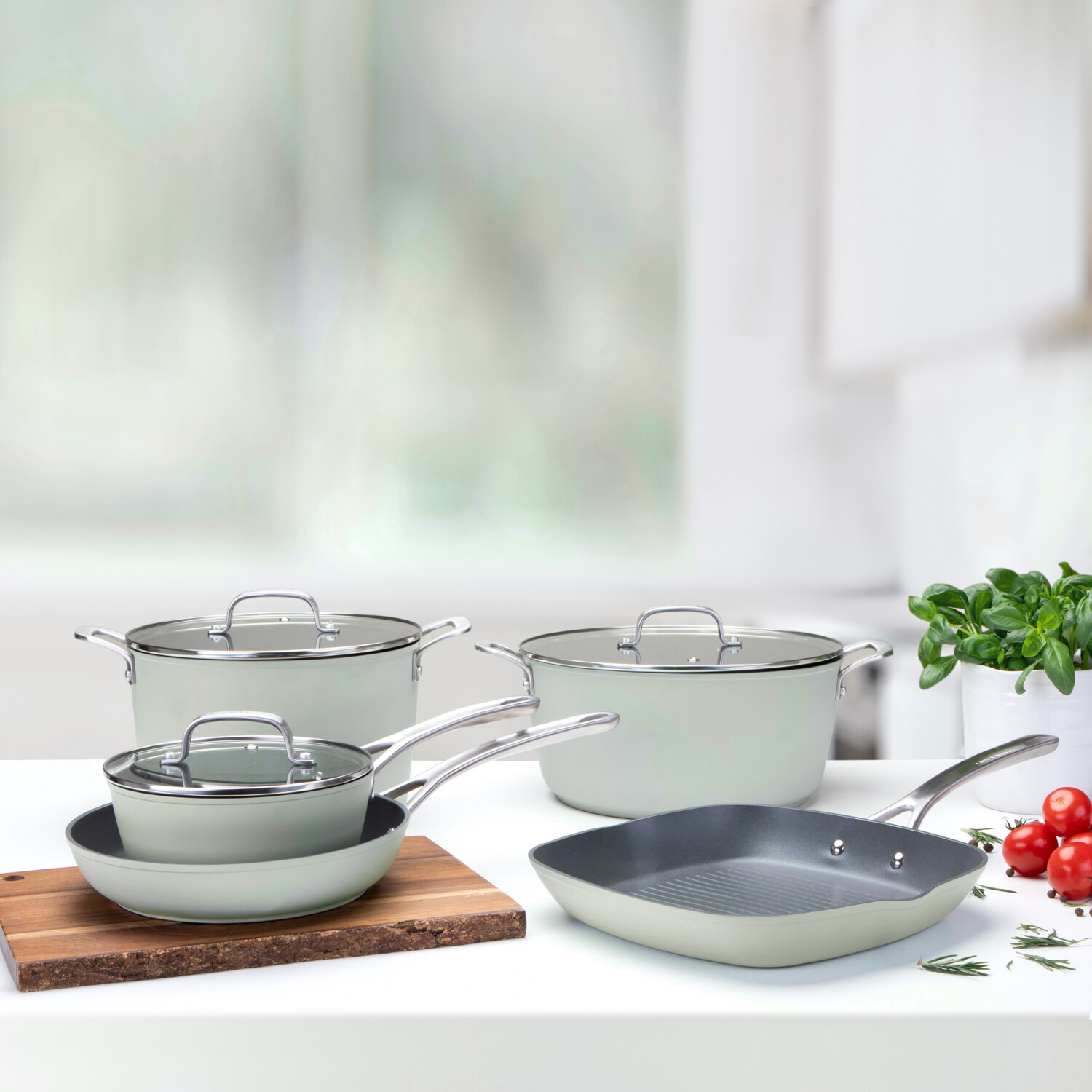 Kochgeschirr-Set auf einem weißen Tisch. Enthält Pfannen, Töpfe mit Deckel. Tomaten, Kräuter und eine Pflanze im Hintergrund. Küchenumgebung.