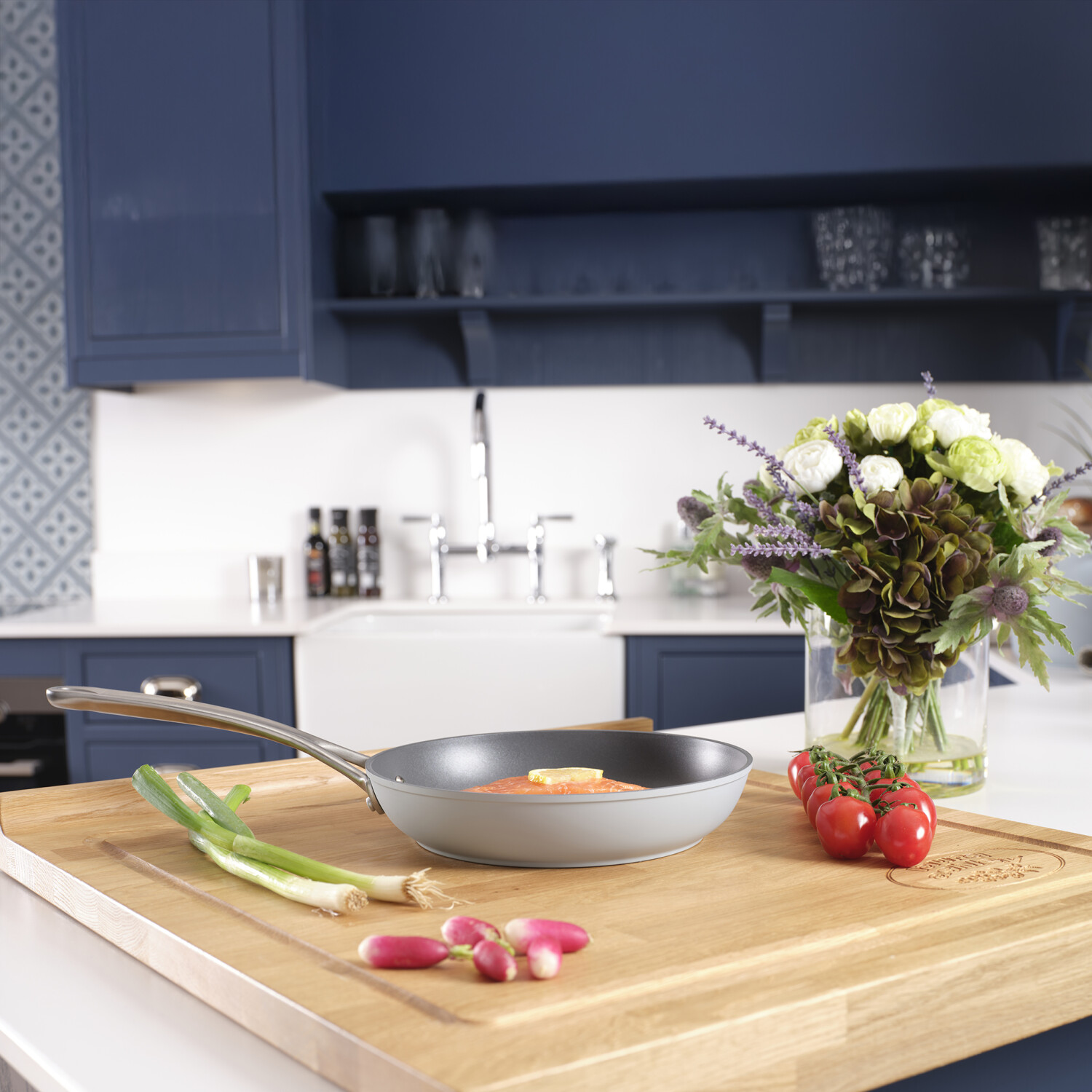 Eine Küche mit einer Pfanne mit Lachs, Gemüse und Blumen auf einem Holzbrett. Dunkelblaue Schränke im Hintergrund.