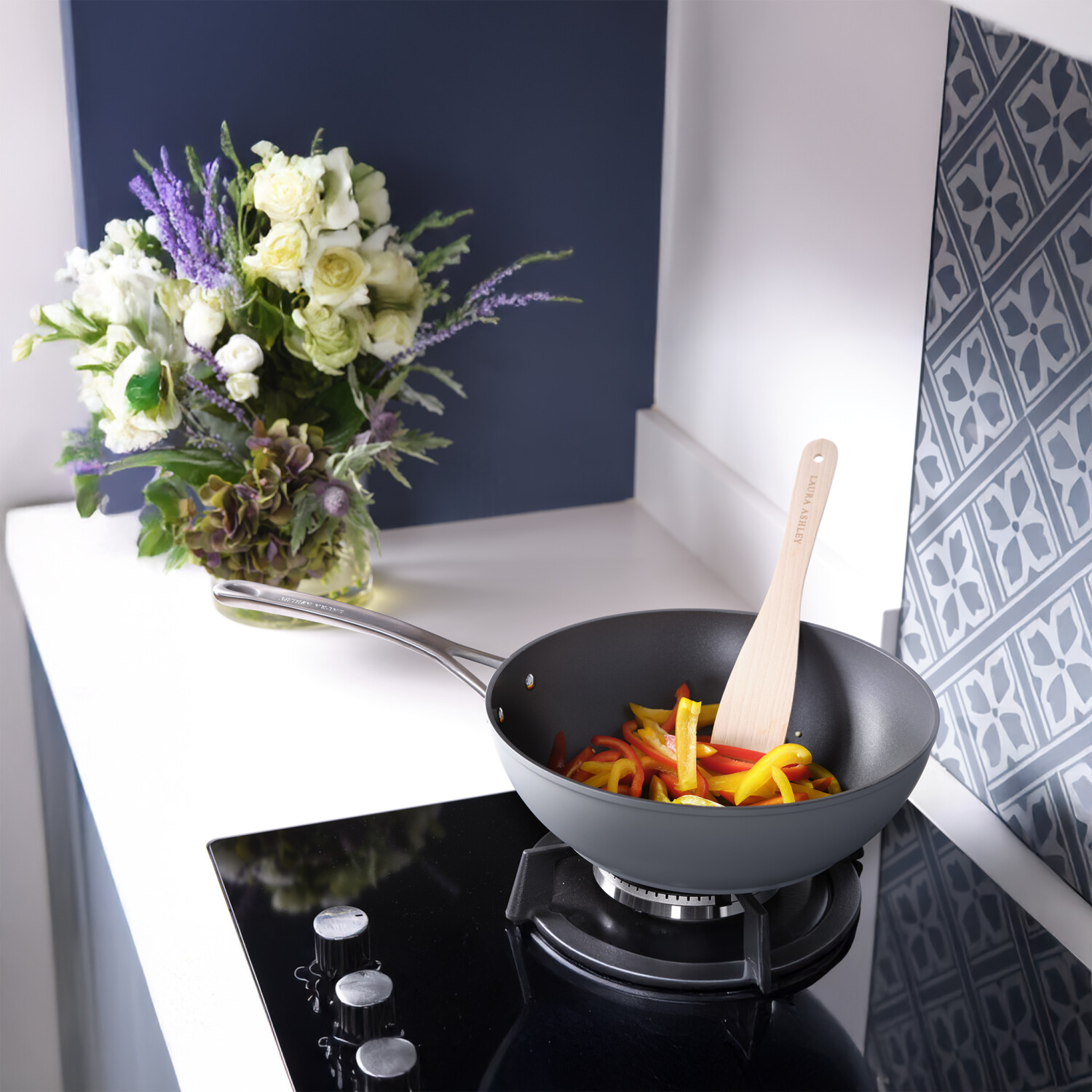 Ein Wok mit Paprika, der mit einem Holzspatel umgerührt wird. Ein Blumenstrauß und eine gemusterte Wand sind im Hintergrund.