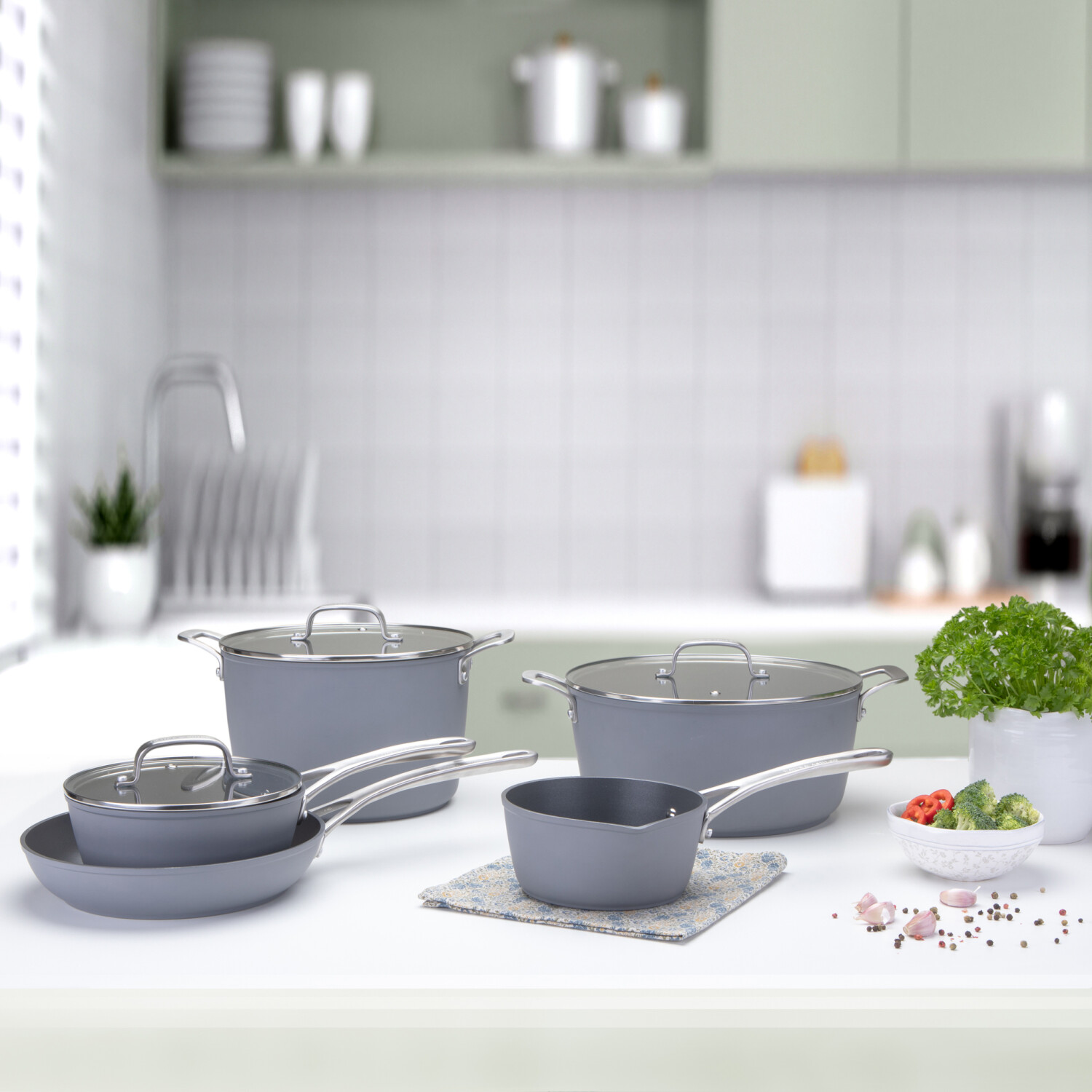 Küchenszene mit grauem Kochgeschirr, Gemüse und Kräutern auf einer weißen Arbeitsplatte. Im Hintergrund eine verschwommene Küche.