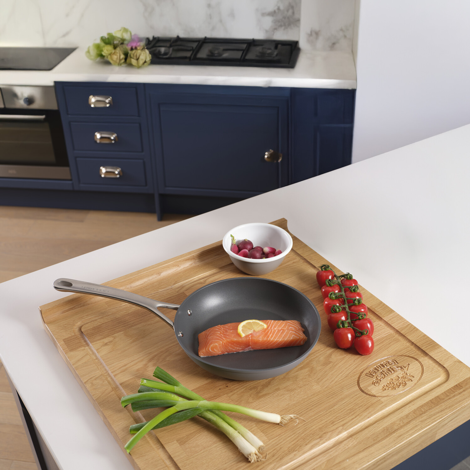 Küchenszene mit Lachs in einer Pfanne, Tomaten, Frühlingszwiebeln und Radieschen auf einem Holzbrett. Hintergrund: blaue Schränke und Herd.