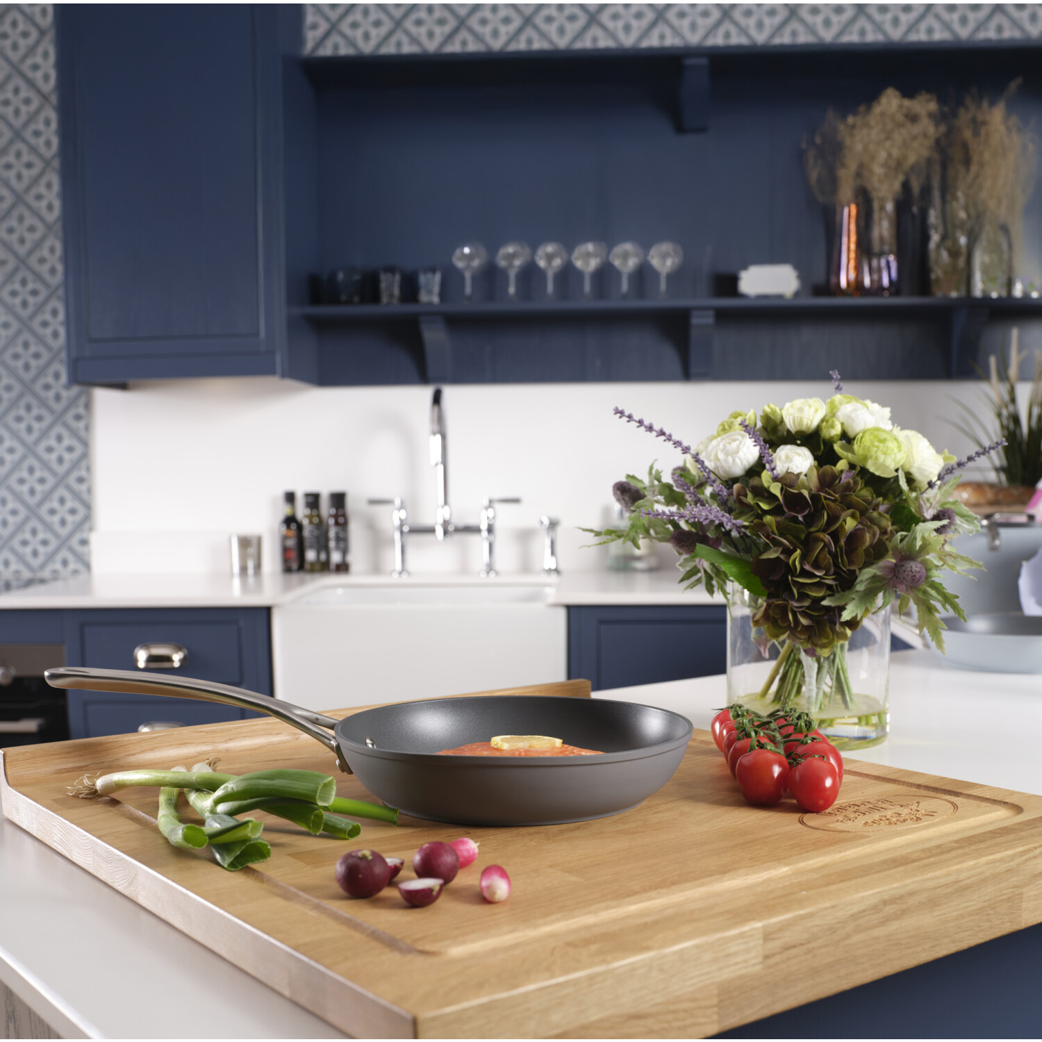Eine Küchenszene mit einer Pfanne auf einem Holzbrett mit Gemüse und Blumen. Der Hintergrund umfasst Schränke, Spüle und Glaswaren.