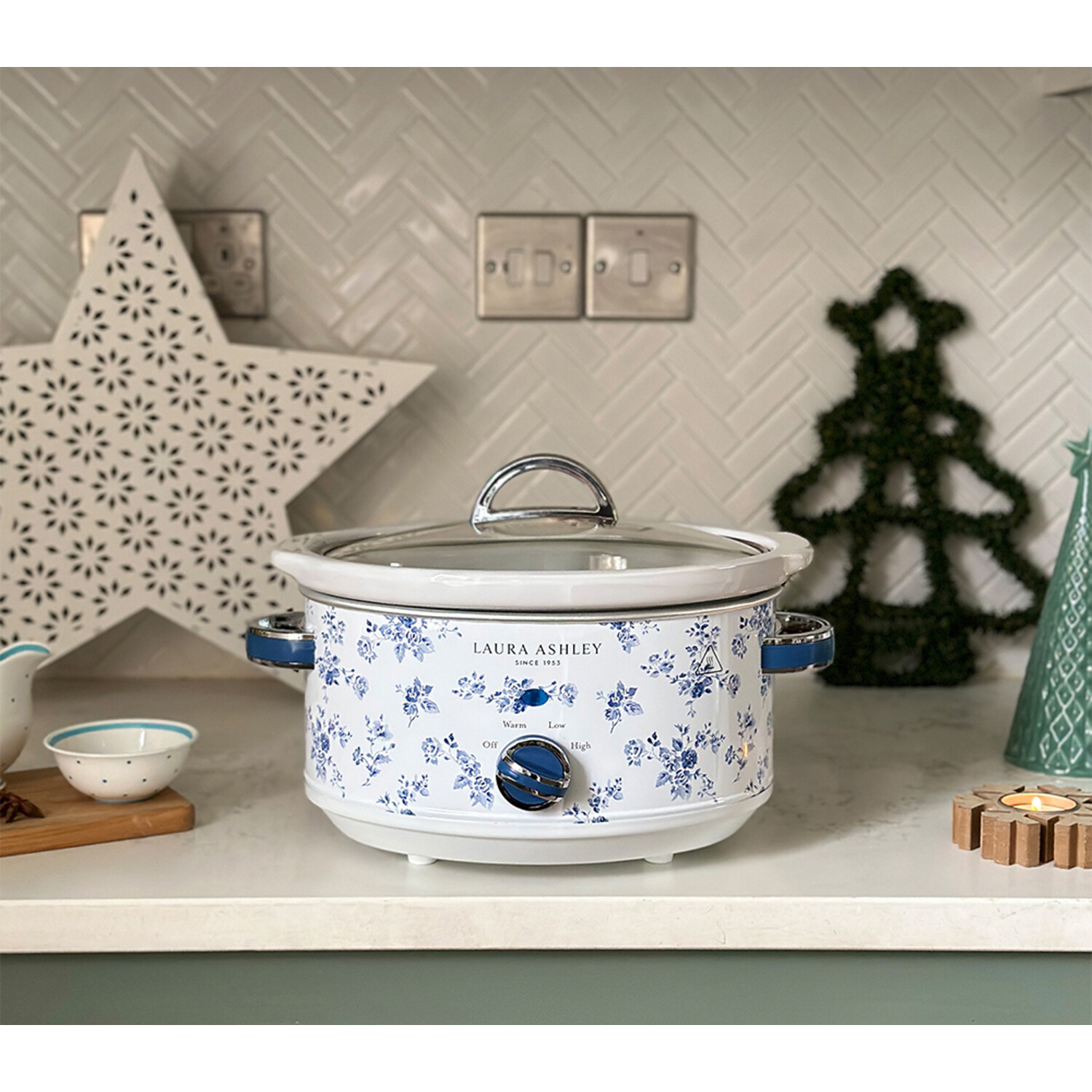 Ein weißer und blauer Blumen-Slowcooker steht auf einer Theke. Ein Stern und ein Weihnachtsbaum sind dahinter. Andere Gegenstände sind im Bild.