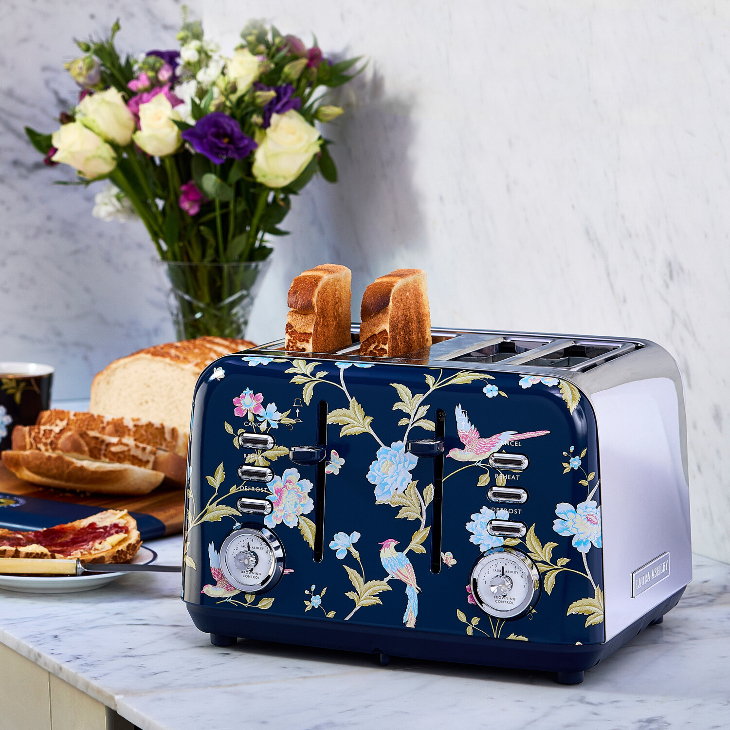 Ein floraler Toaster mit Toastbrot. Daneben Brot, Marmelade und Blumen in einer Vase auf einer Marmoroberfläche.