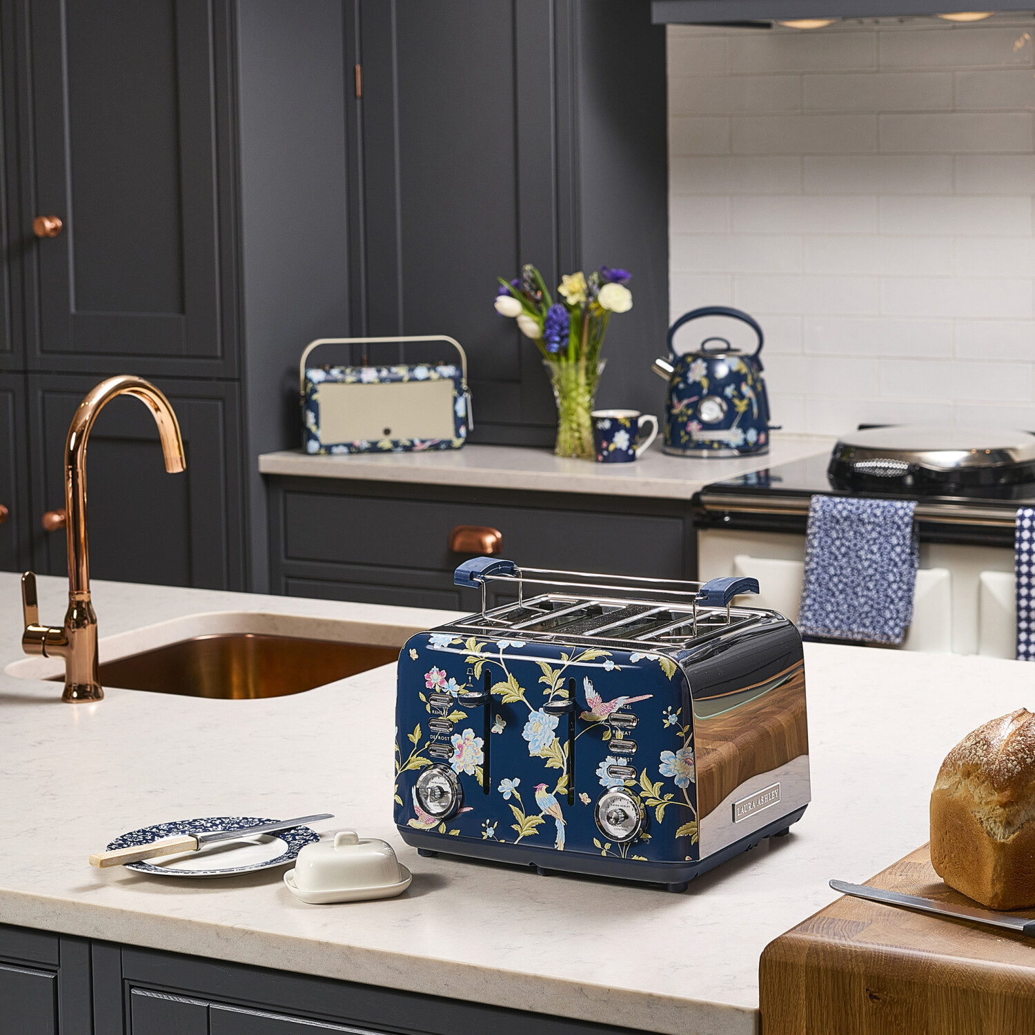 Eine Küchenszene mit einem floralen Toaster, Wasserkocher und Zubehör auf einer Arbeitsplatte. Ein Brotlaib und ein Kupferhahn sind ebenfalls vorhanden.