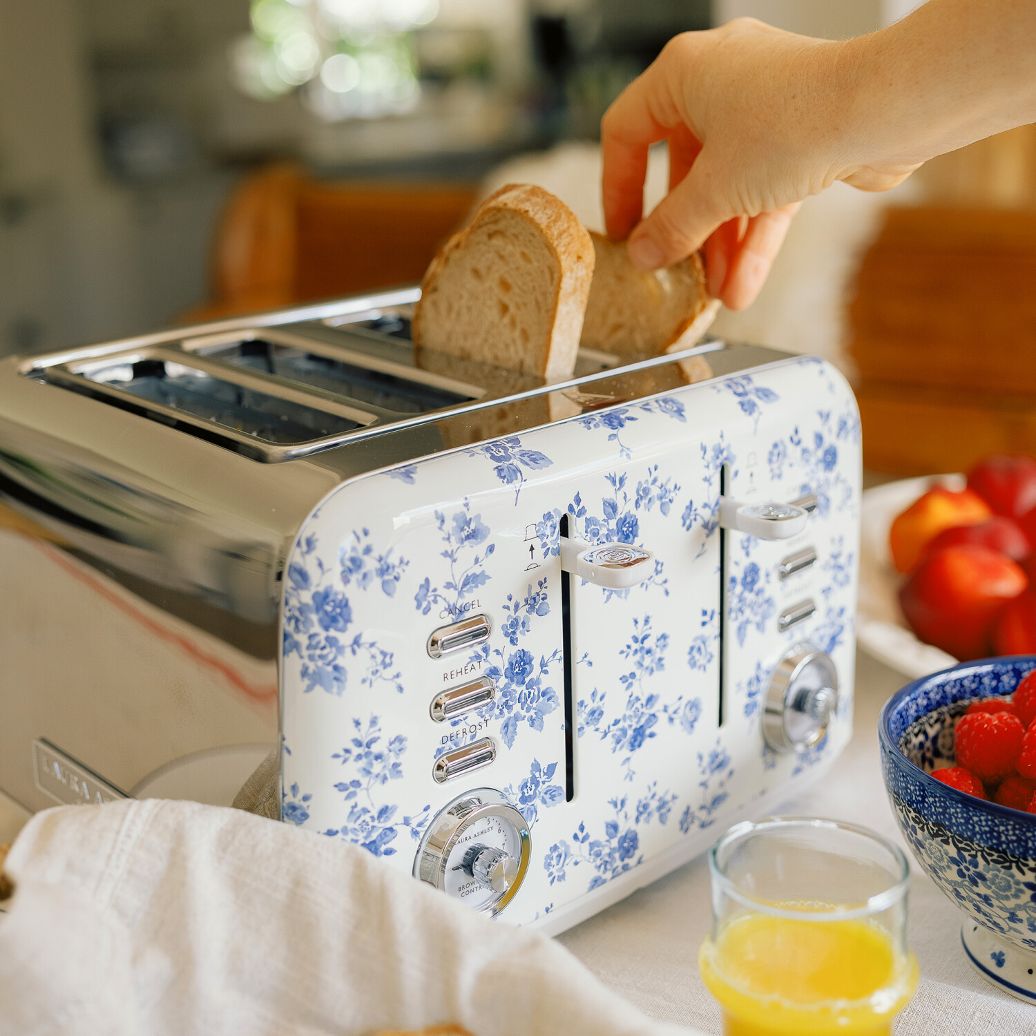 Eine Hand legt Brot in einen floralen Toaster. Daneben liegen Früchte, Saft und eine Schüssel mit Beeren auf einer weißen Tischdecke.