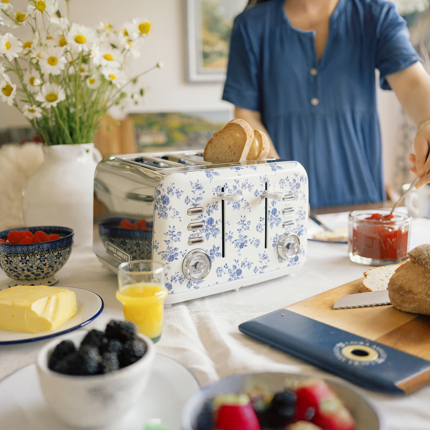 Eine Frühstücksszene mit einem floralen Toaster, Brot, Butter, Beeren, Saft und einer Person, die Marmelade auf Toast streicht.