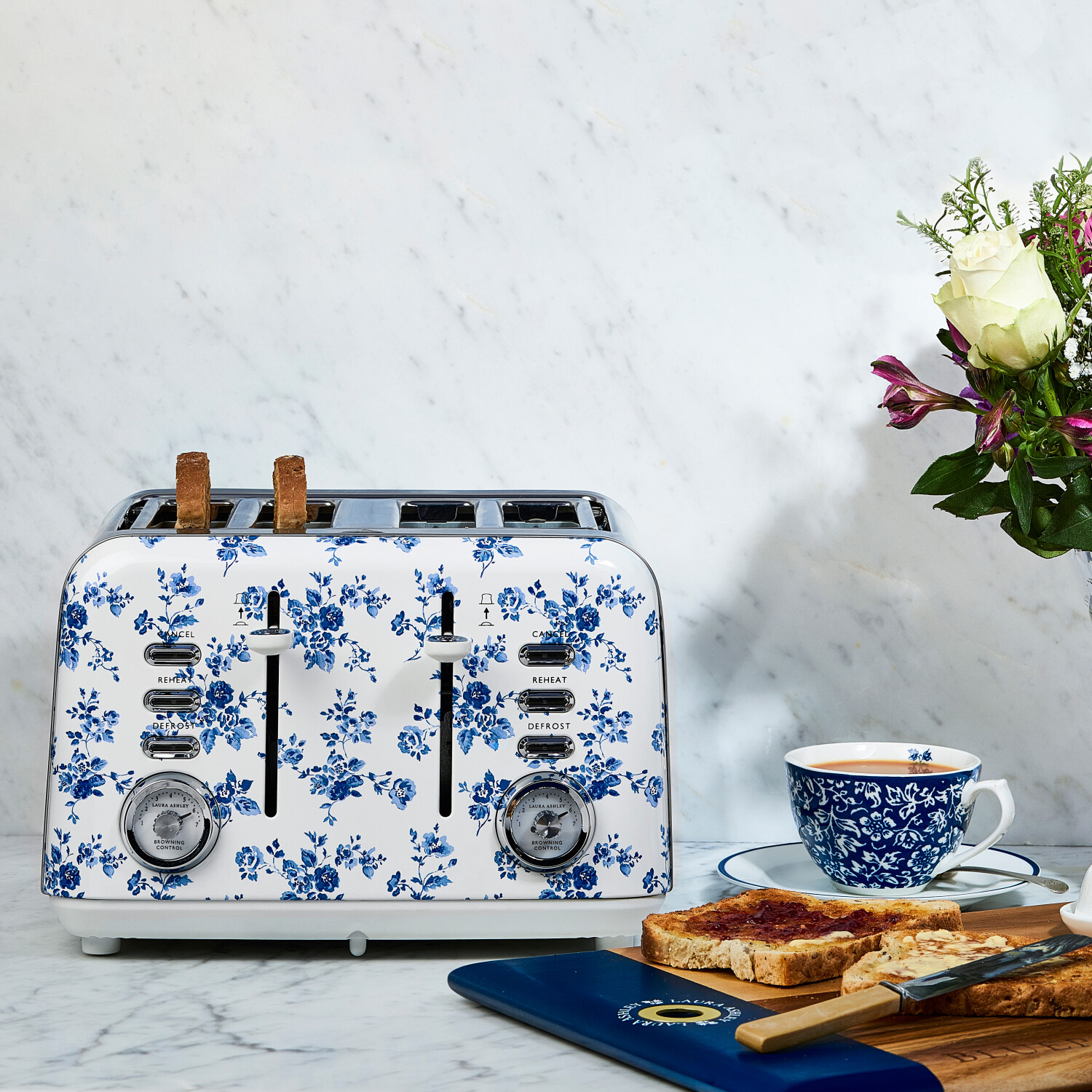 Ein floraler Toaster mit Toast, eine Tasse Tee und Blumen stehen auf einer Marmoroberfläche. Toast hat Marmelade und Butter.
