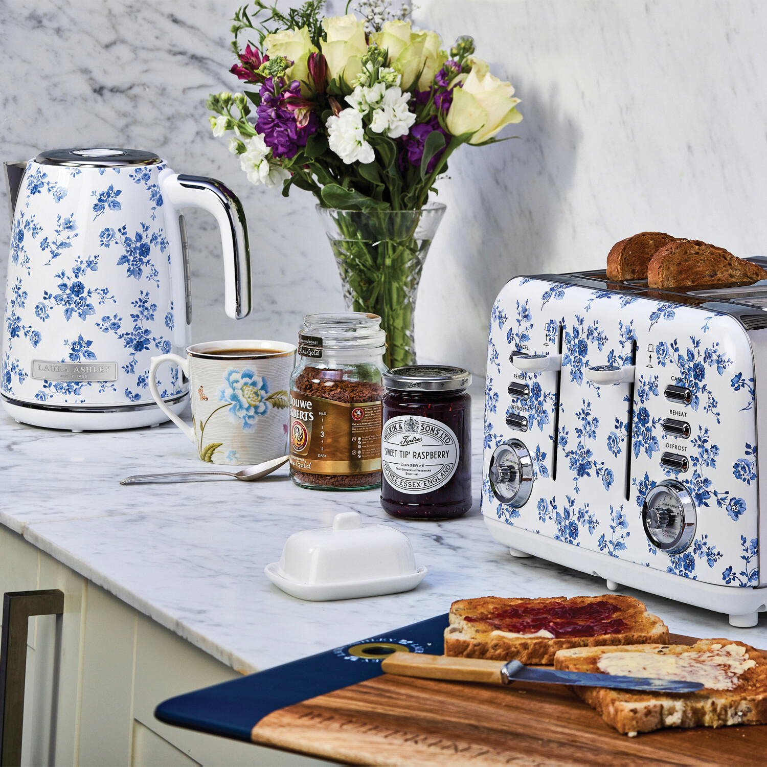 Eine Küchenszene mit floral gemusterten Geräten, Brot, Toast mit Marmelade, Blumen und Gläsern auf einer Marmorarbeitsplatte.