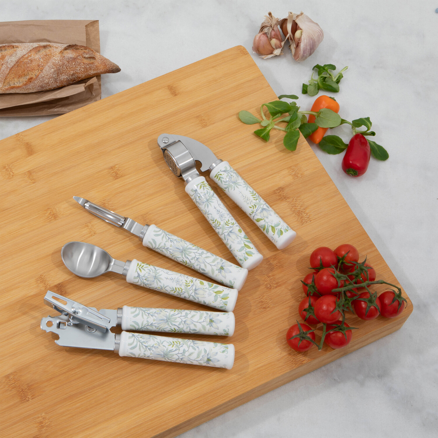 Ein hölzernes Schneidebrett mit Küchenutensilien, Tomaten, Knoblauch und Brot. Werkzeuge umfassen eine Knoblauchpresse und einen Dosenöffner.