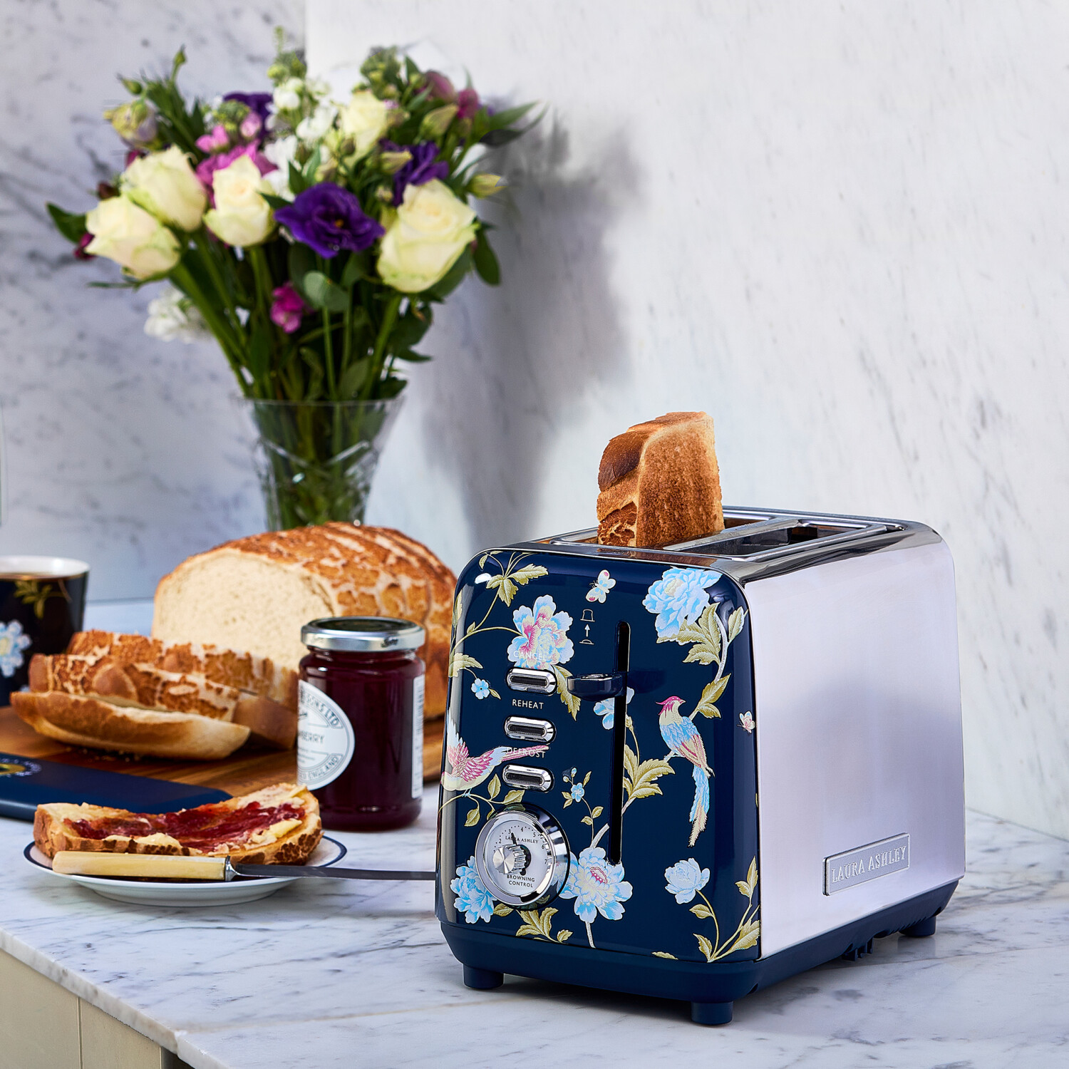 Ein floraler Toaster mit Toast, Brot, Marmelade, Blumen und einem Teller Toast mit Marmelade stehen auf einer Marmoroberfläche.