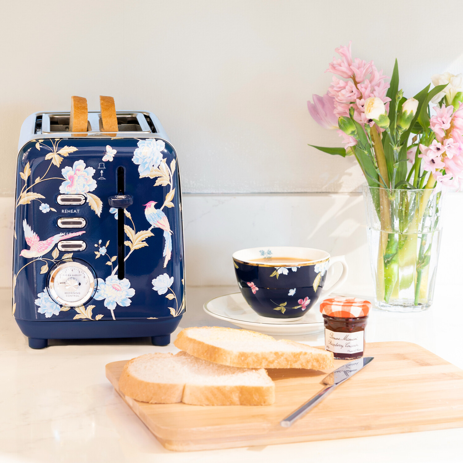 Ein marineblauer, floraler Toaster mit Toast, Tasse Tee, Blumen und Marmeladenglas auf einem Holzbrett. Helle, saubere und einladende Frühstücksszene.