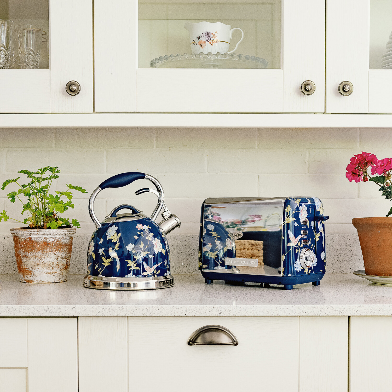 Eine Küchenszene mit einem floralen Wasserkocher, Toaster und Pflanzen. Eine Teetasse ist im Schrank sichtbar, der Stil ist floral.