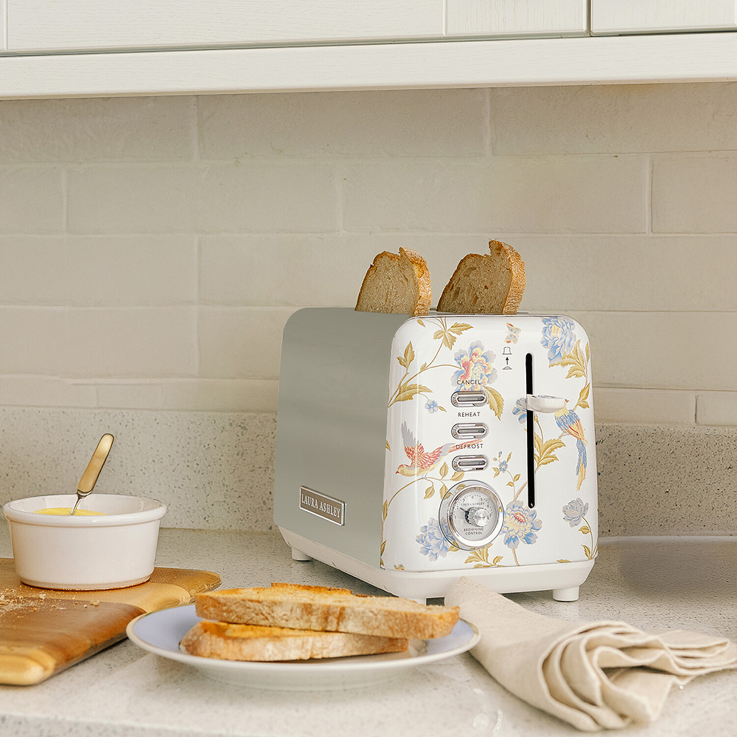 Ein floraler Toaster mit Brot, Toast auf einem Teller, eine Schüssel mit Butter und eine Serviette auf einer Theke.
