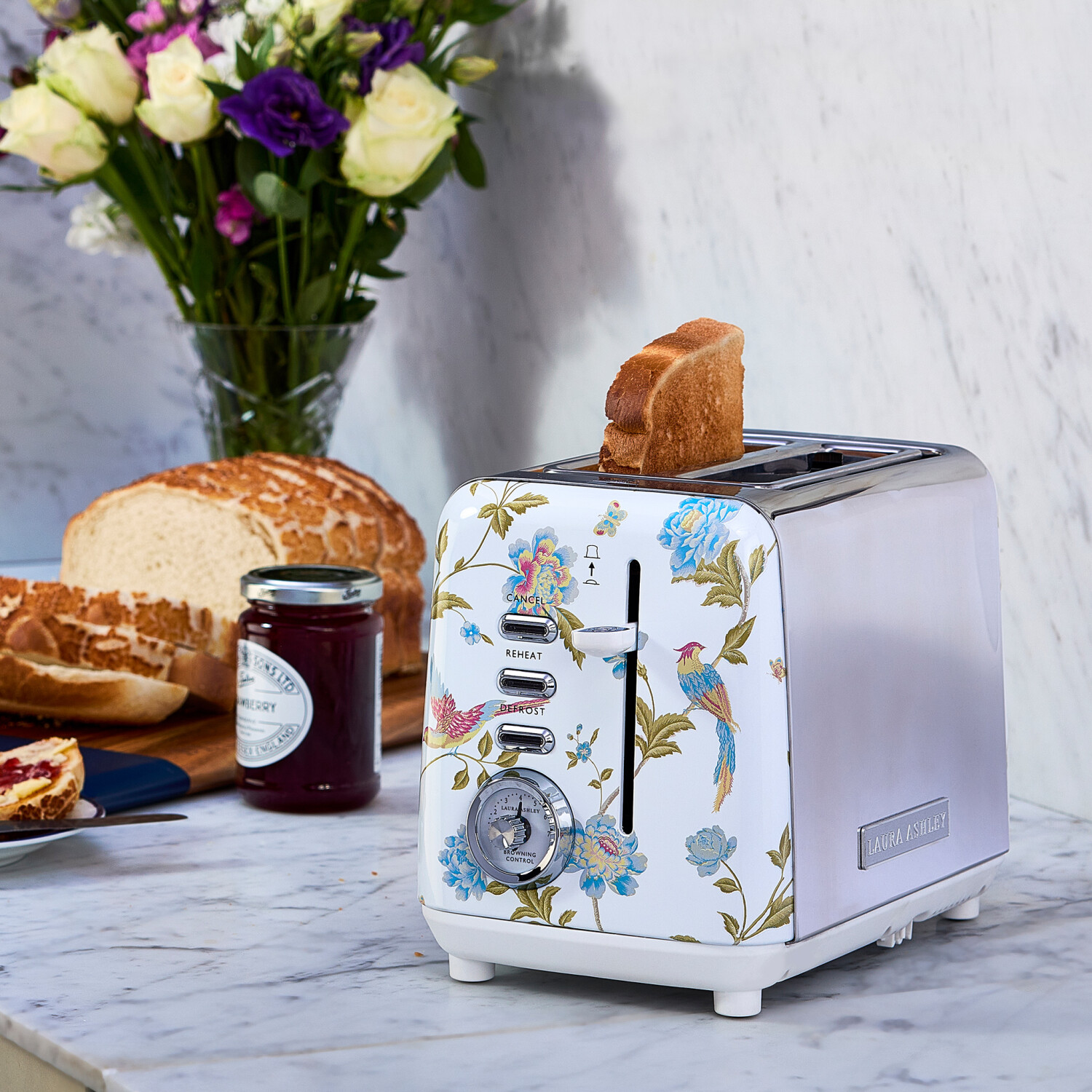 Ein floraler Toaster mit herausspringendem Toast, Brot, Marmelade und Blumen auf einer Marmortheke. Eine Scheibe Toast steckt im Toaster.