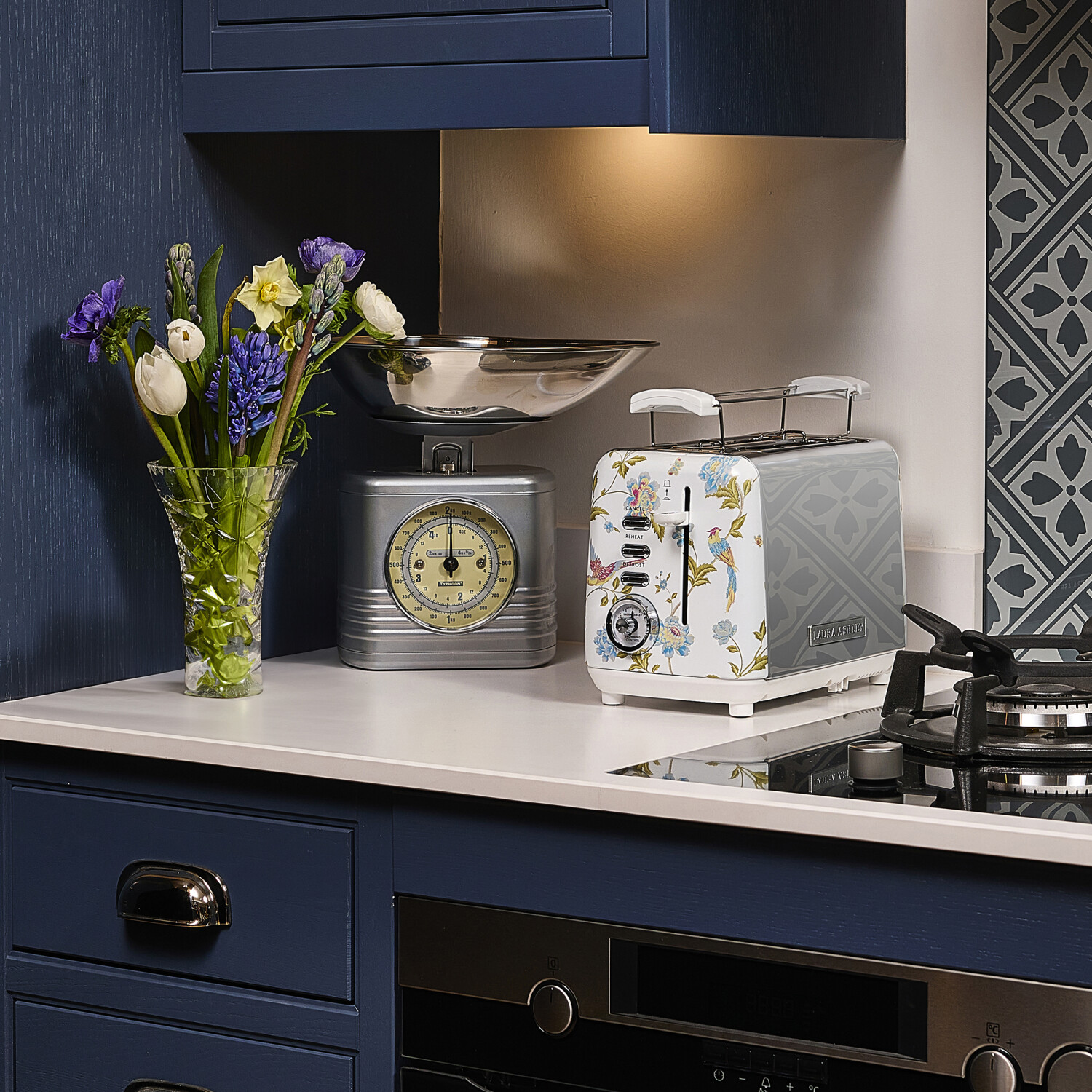 Küchenszene mit Blumen, Waage und Toaster. Blaue Schränke und gemusterter Fliesenspiegel. Weiße Arbeitsplatte.