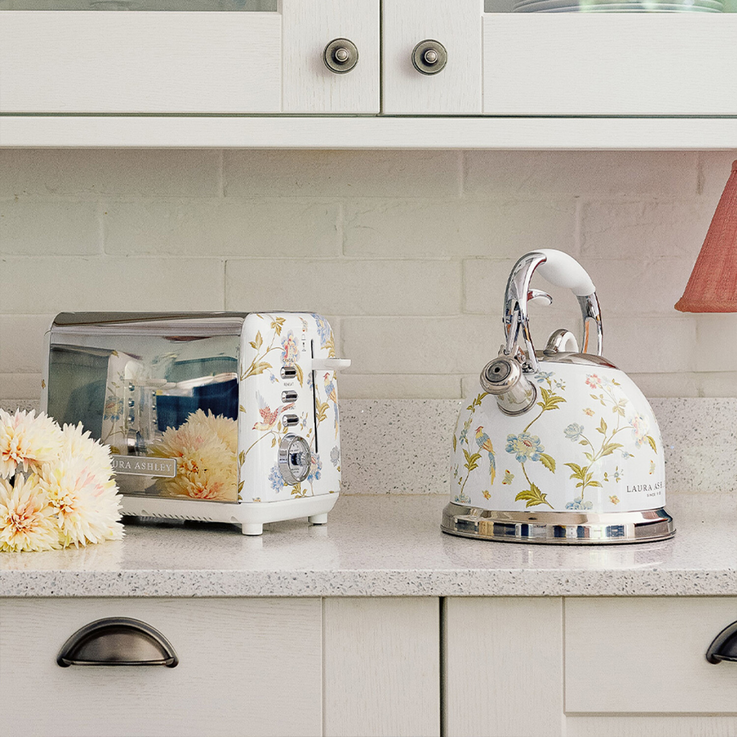 Eine Küchenszene mit einem floralen Toaster und Wasserkocher auf einer Arbeitsplatte. Weiße Schränke und eine Ziegelmauer sind im Hintergrund.