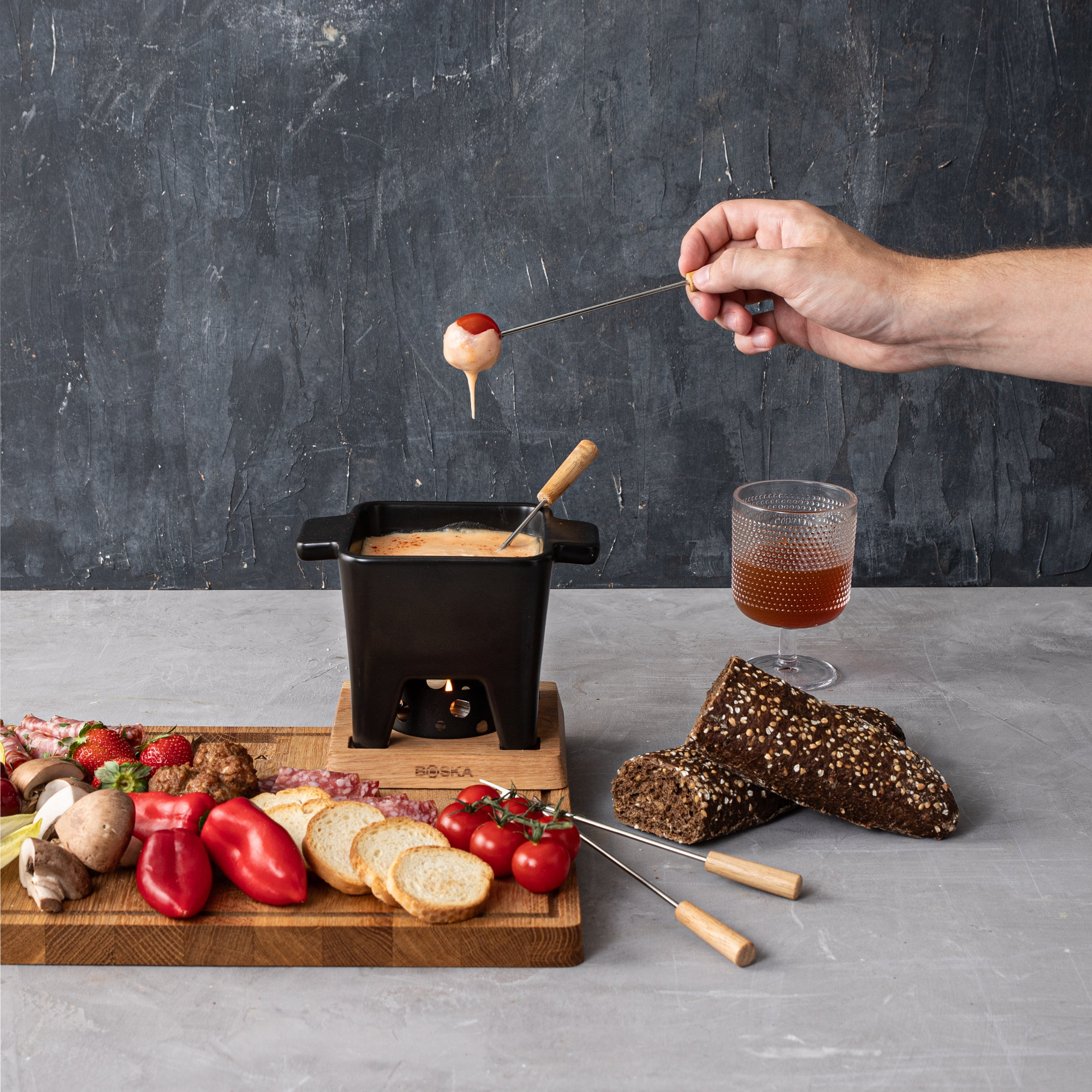 A fondue setup with a hand holding a cheese-covered food. Includes bread, vegetables, and a drink.