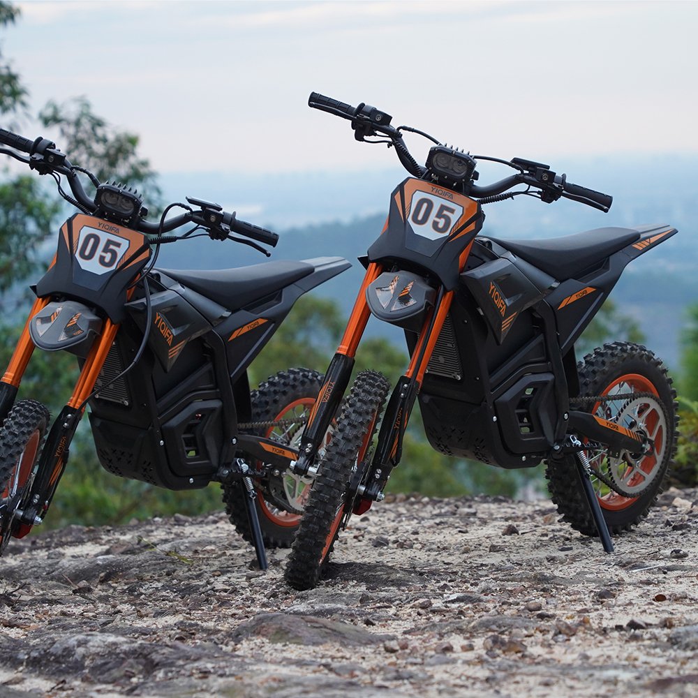 Zwei schwarze und orangefarbene Elektro-Dirtbikes mit der Nummer 05 vorne, geparkt auf felsigem Untergrund, mit Bäumen und einem bewölkten Himmel.