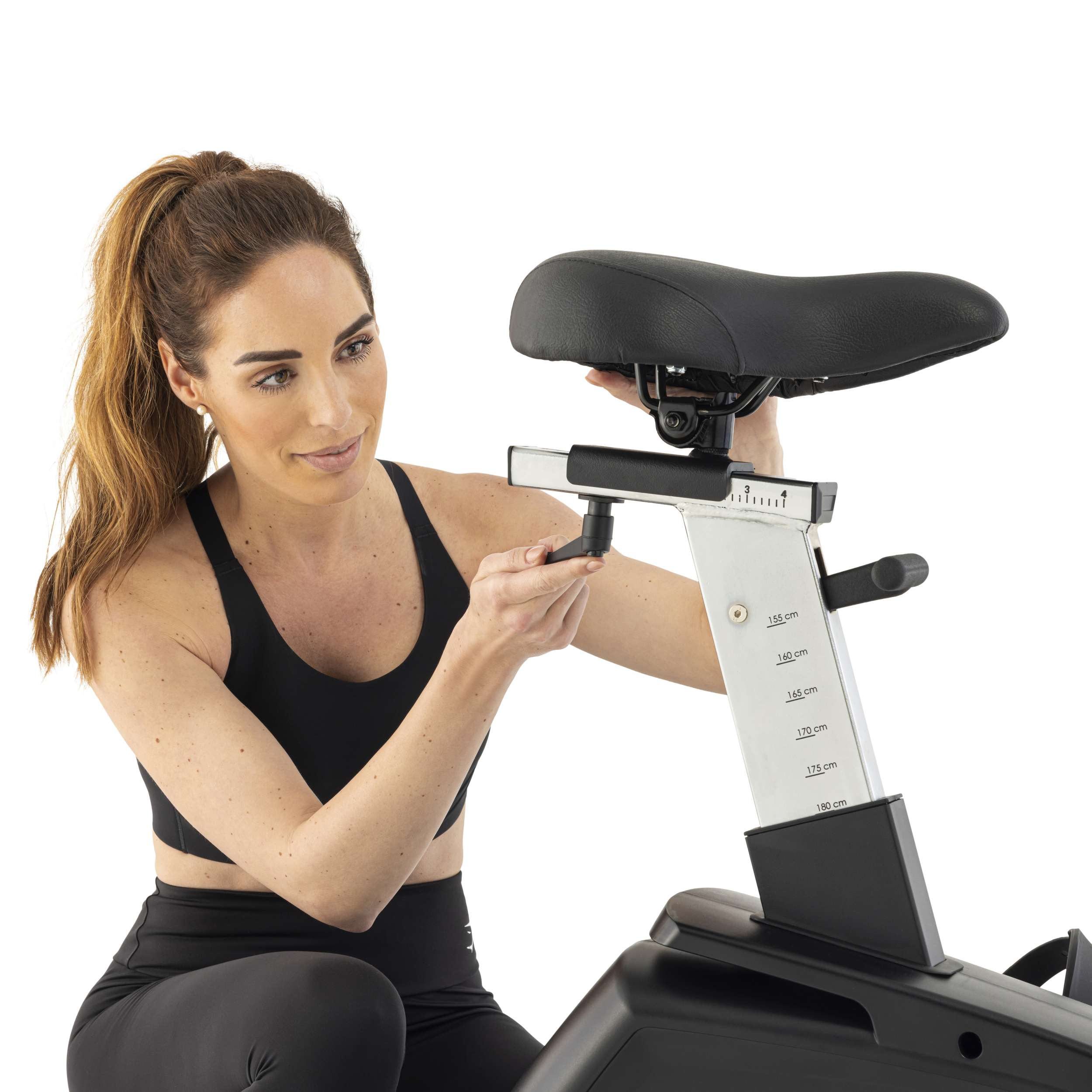 Vrouw in zwarte sportkleding die de zitting van een hometrainer verstelt, geïsoleerd op witte achtergrond.