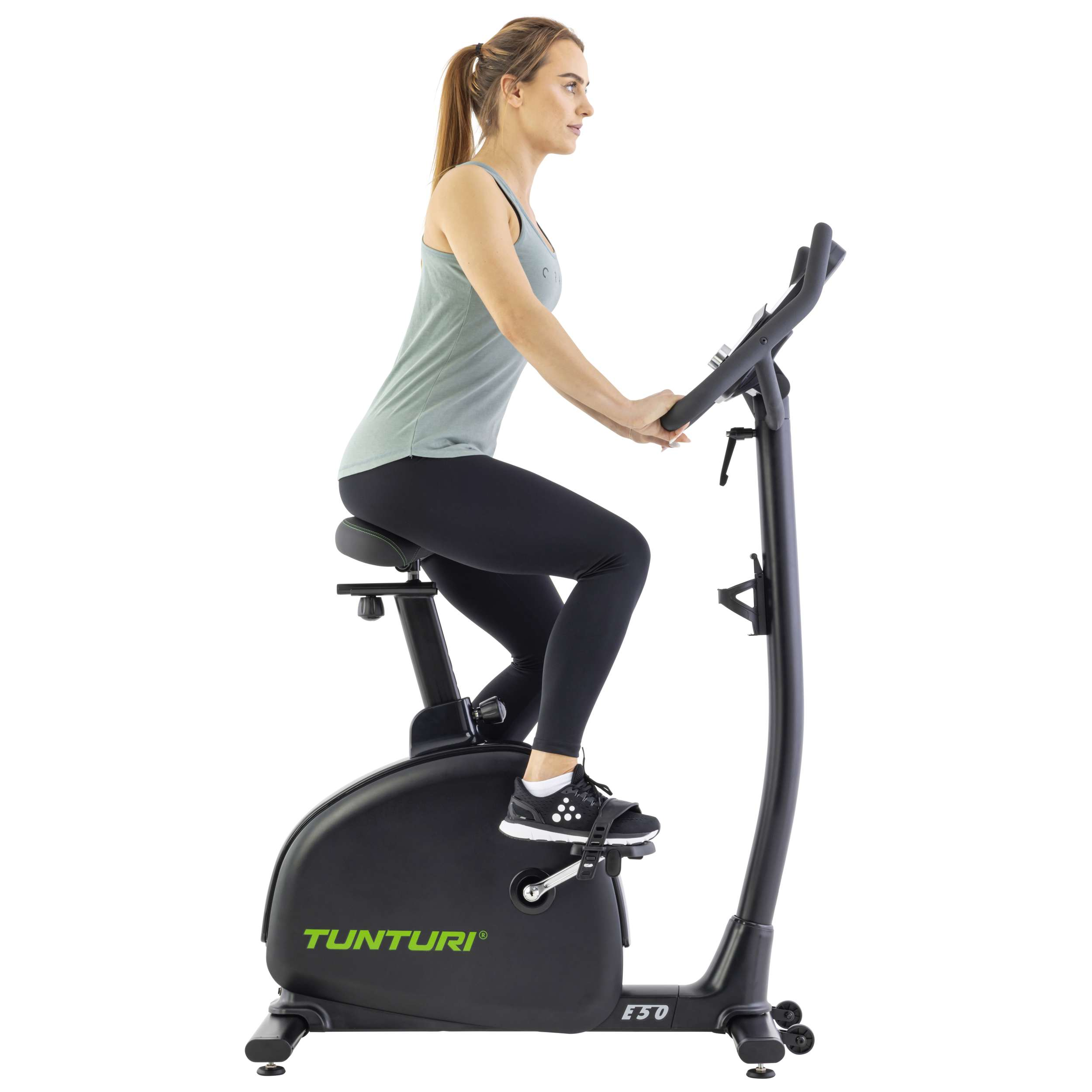 Een vrouw sport op een hometrainer. Ze draagt een zwarte legging en een groene top. De fiets is zwart met groene accenten.