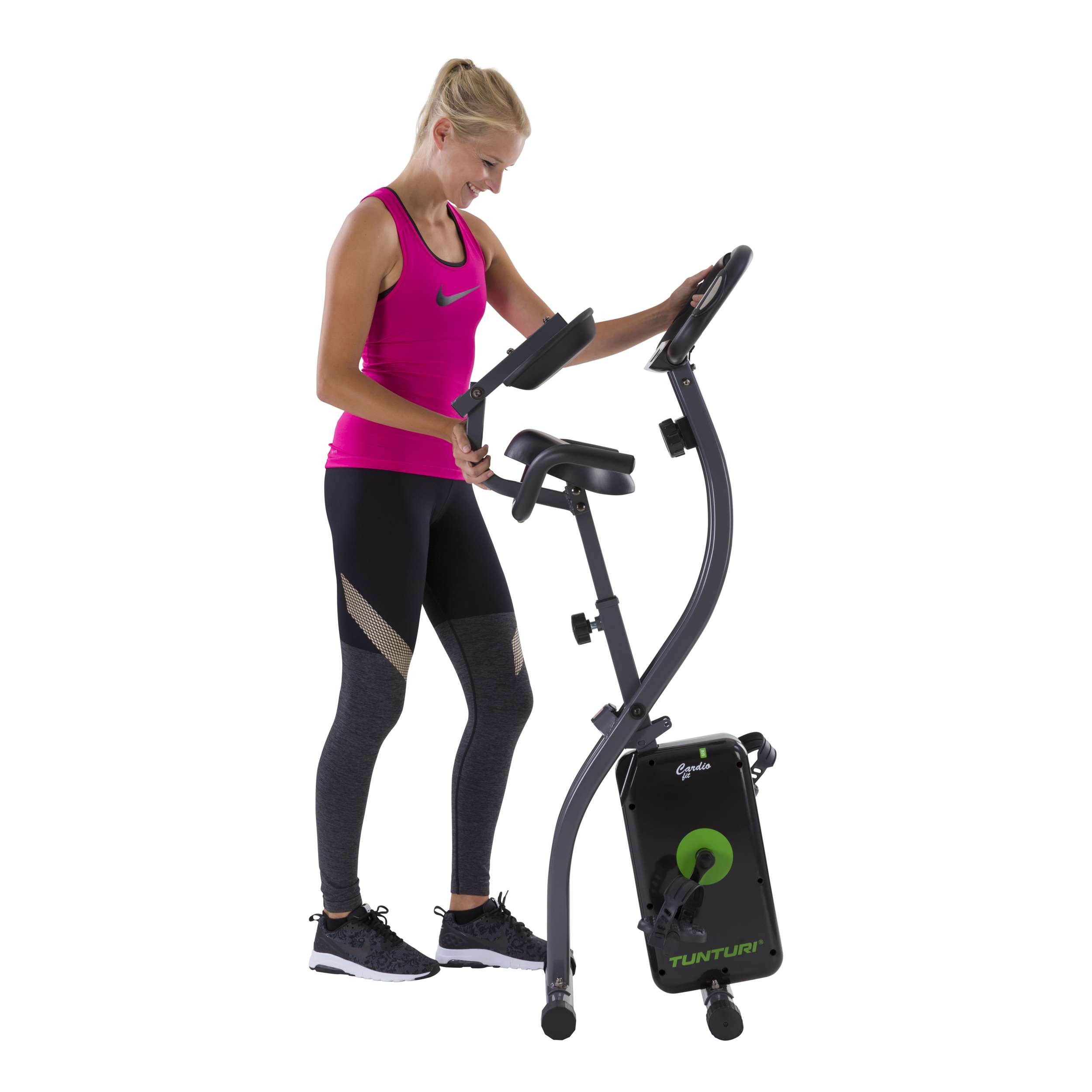 Vrouw in roze tanktop die een hometrainer gebruikt. Ze lacht. Zwarte legging en schoenen. Witte achtergrond.